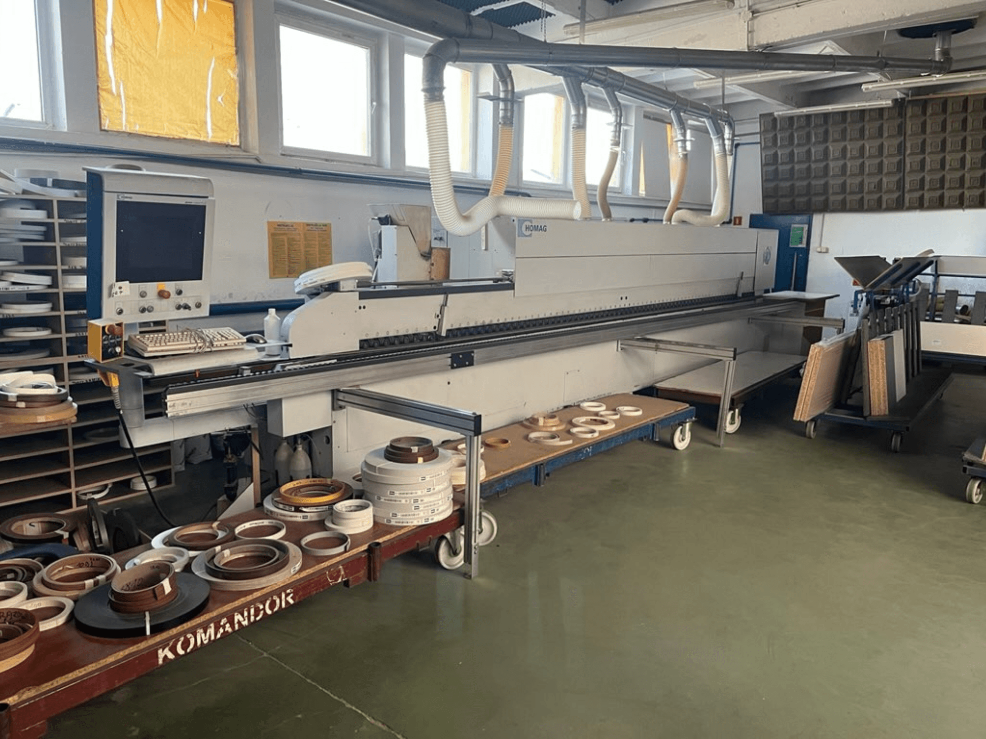 HOMAG-reunanauhakone työpajassa, jossa on hallintalaitteet ja erilaisia reunanauhamateriaaleja sinisellä kärryllä. Sivukuva.