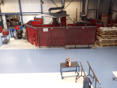 Teollinen työtila, jossa on punaisia hitsausverhoja, koneita ja kuormalavoja; ylhäältä päin näkymä, jossa näkyy ulkoasu ja laitteet.