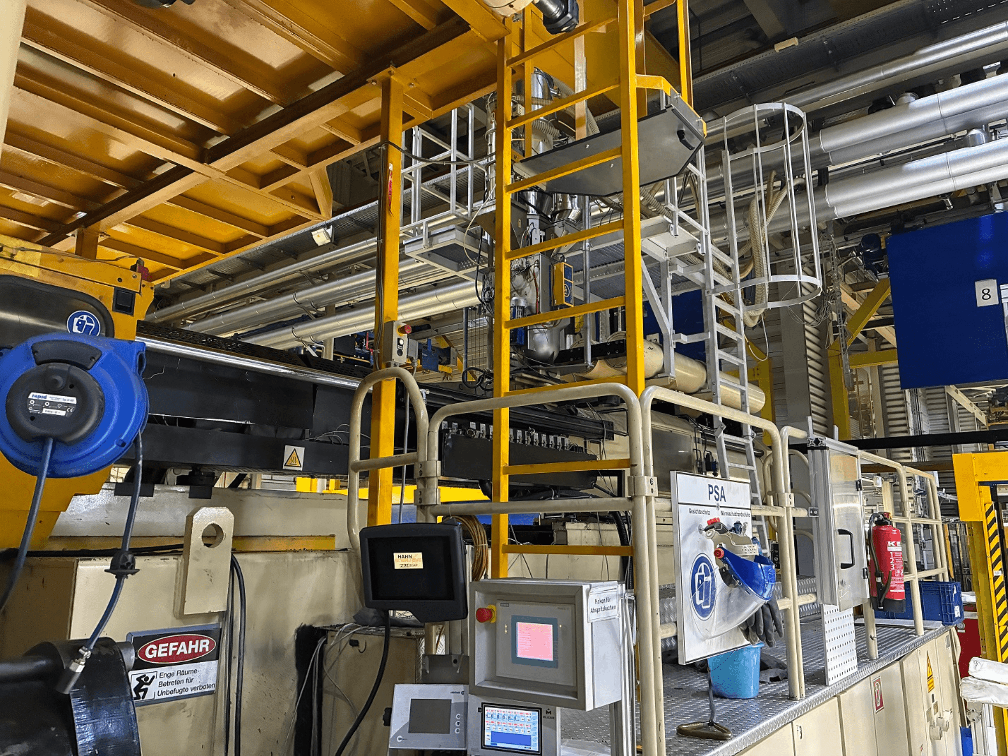 Teollisuuskoneiden ohjauspaneeli, jossa on siniset turvalaitteet ja keltaiset telineet, sivukuva, jossa näkyy putket ja varoitusmerkit.