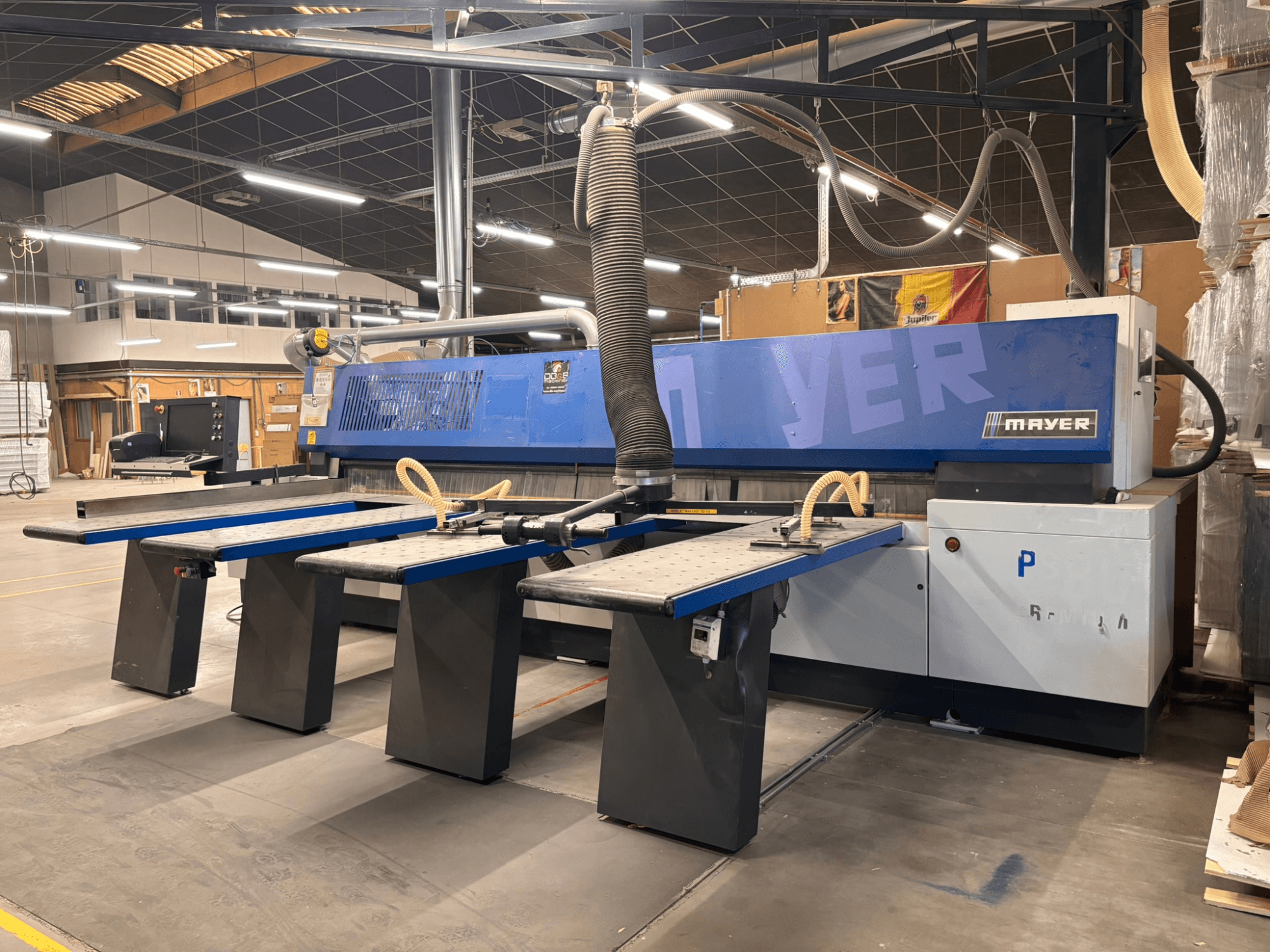 Mayer CNC-kone sinisillä paneeleilla, etukuva; siinä on useita työpöytiä ja pölynpoistojärjestelmä puuntyöstöä varten.