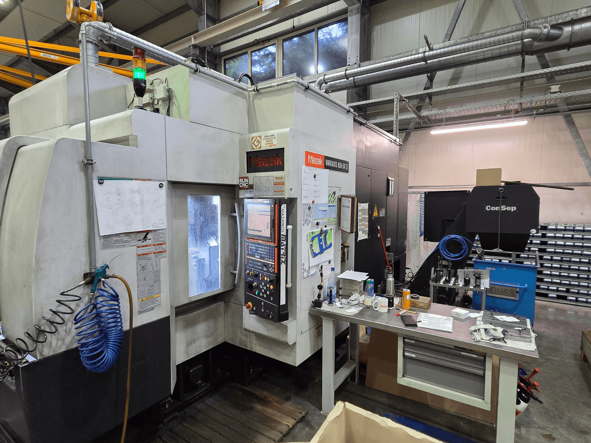 Mazak Variaxis 630-5X CNC-kone työpajassa, etukuva, jossa ohjauspaneeli, letku ja tarvikkeet sivupöydällä.