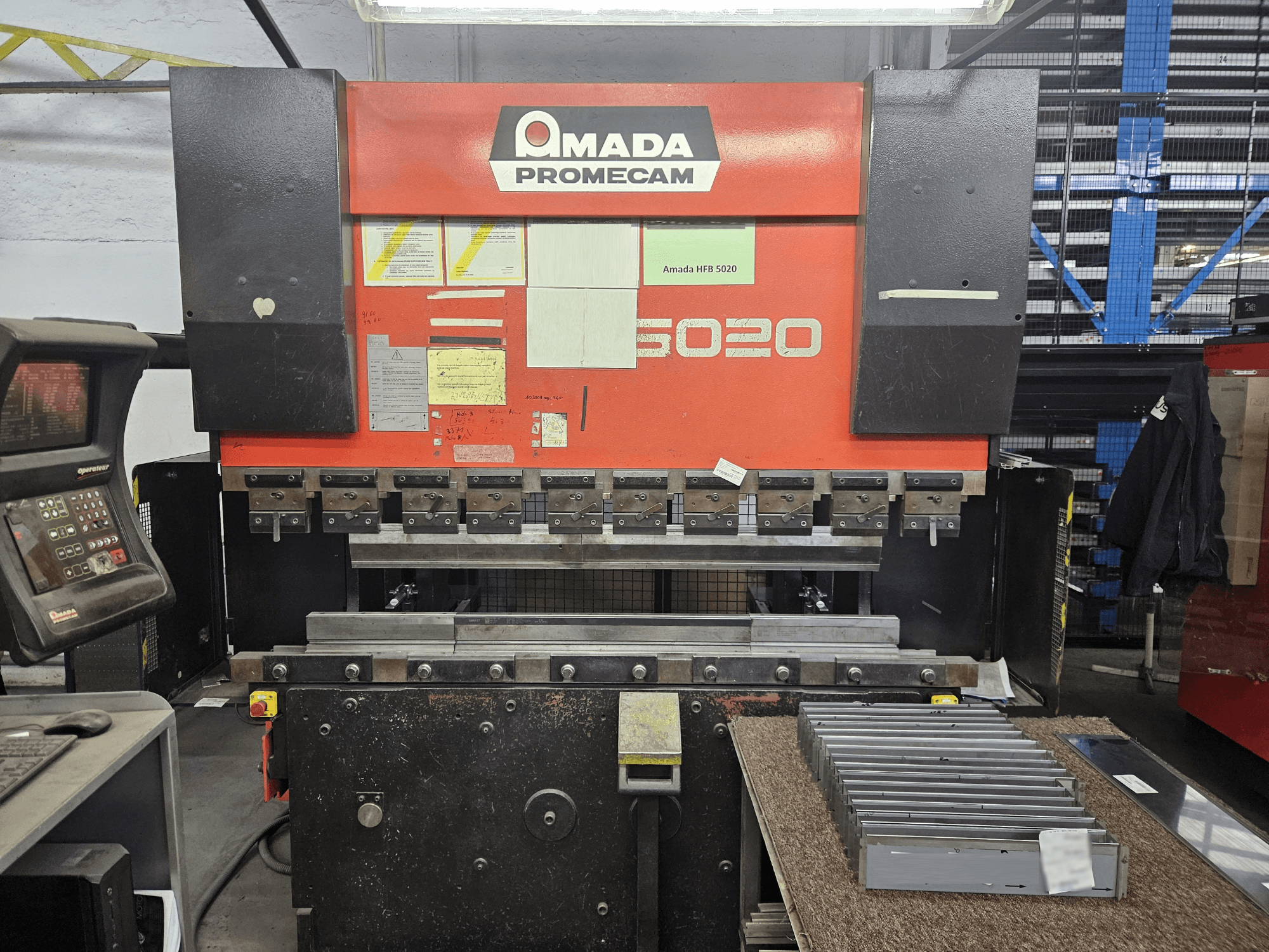 Amada HFBO 5020 -puristimen etukuva, jossa on ohjauspaneeli ja pinottuja metalliosia työtasolla.