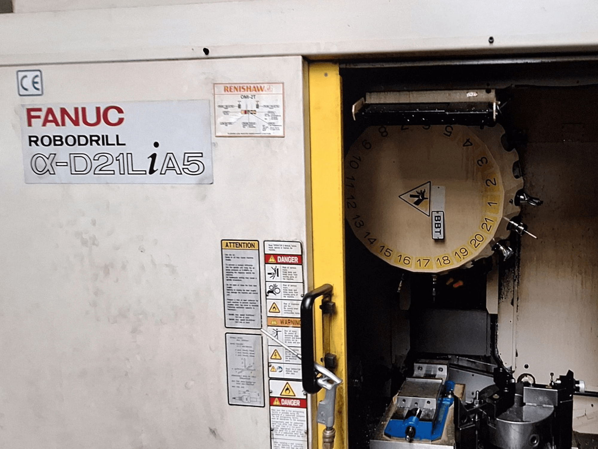 FANUC Robodrill α-D21LiA5, etukuva. Näyttää työkalukarusellin ja työstöalueen, jossa on kiinnittimet ja varoitukset ovessa.