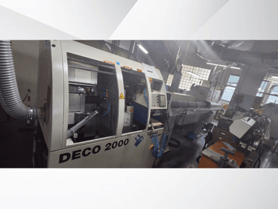 DECO 2000 CNC-kone työpajassa, etukuva, jossa näkyy hallintalaitteet ja oikealla pitkä työkappaleen asetelma.