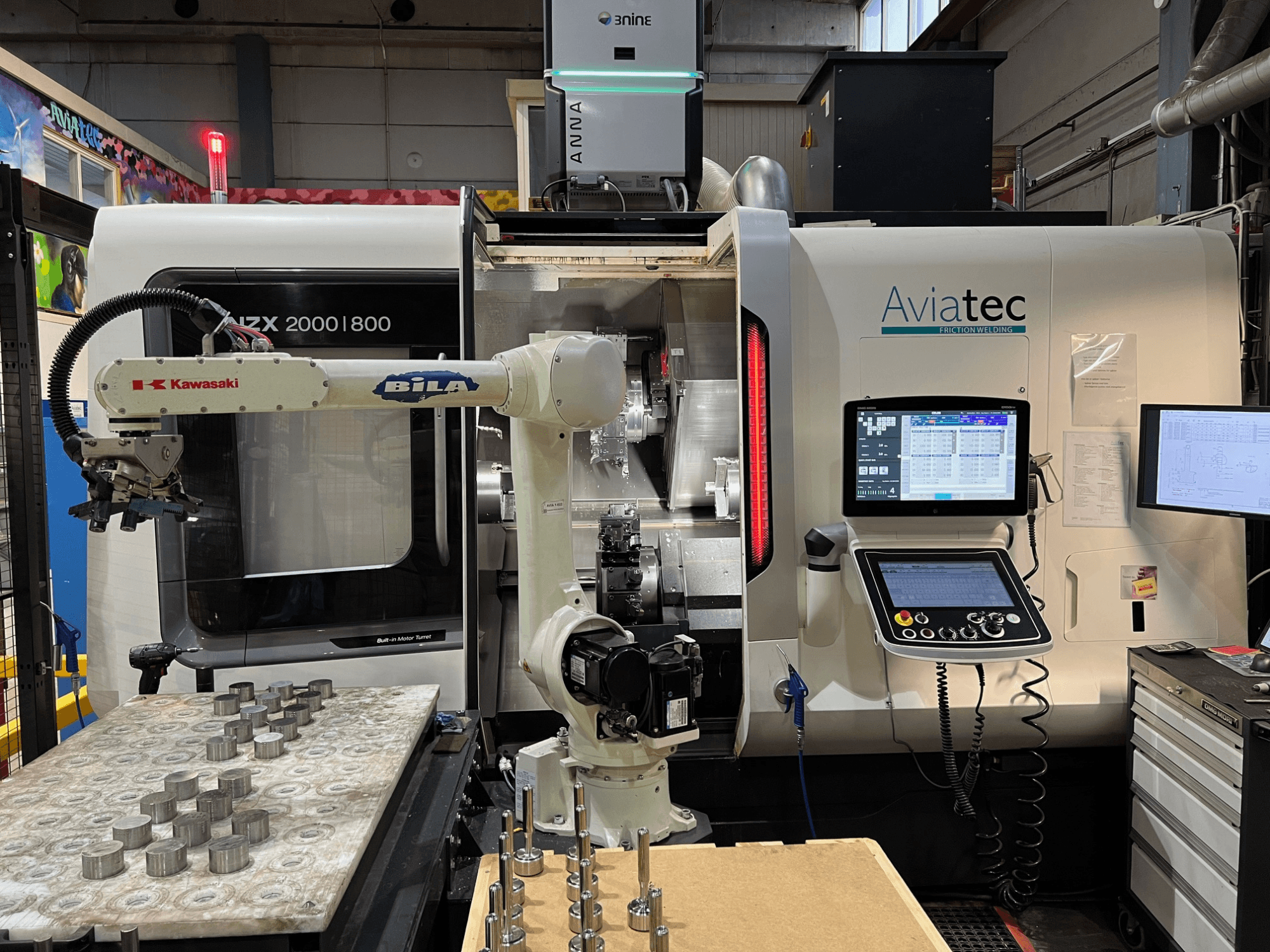 DMG MORI NZX 2000/800SY -vaakasuora sorvi, jossa on Kawasaki-robottivarsi, ohjauspaneeli ja metalliosat työpöydällä.