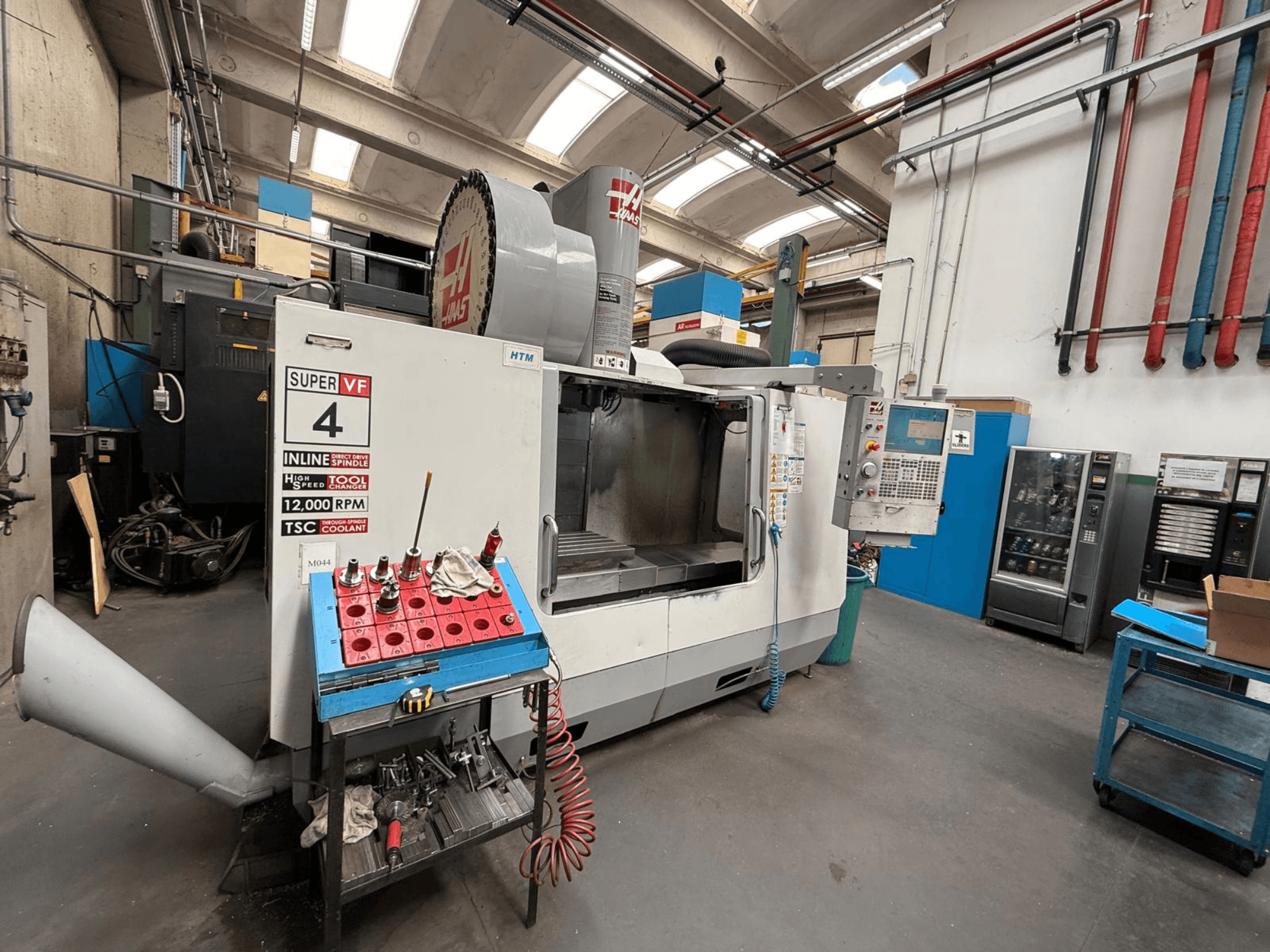 HAAS Super VF 4 CNC-jyrsinkone, jossa on ohjauspaneeli, 12 000 kierrosta minuutissa pyörivä kara ja työkalut sinisessä säilytyshyllyssä, sivukuva.