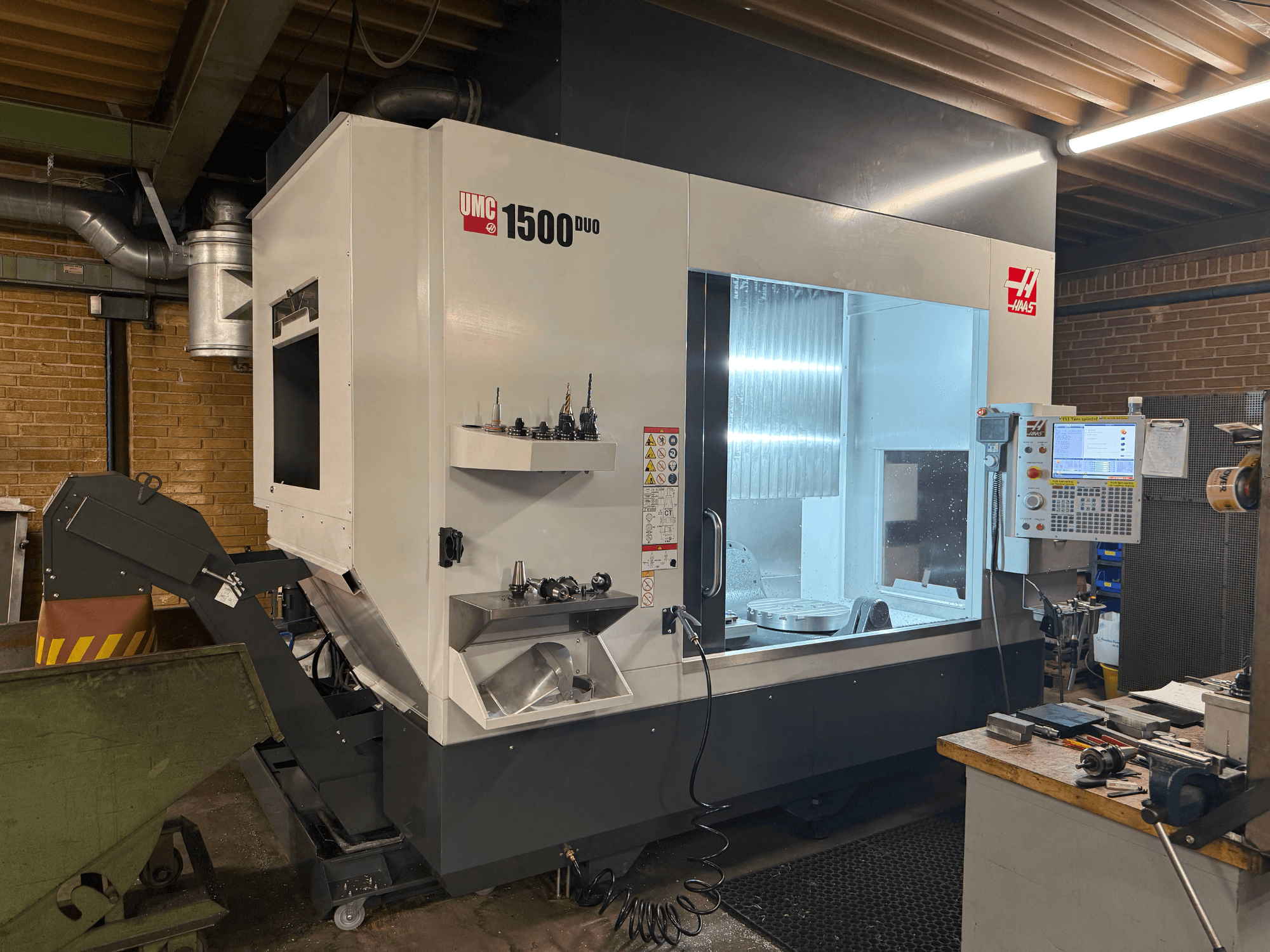 Haas UMC 1500DUO CNC-kone, etukuva, jossa on suuri ikkuna, ohjauspaneeli ja hyllyllä olevat työkalupidikkeet.