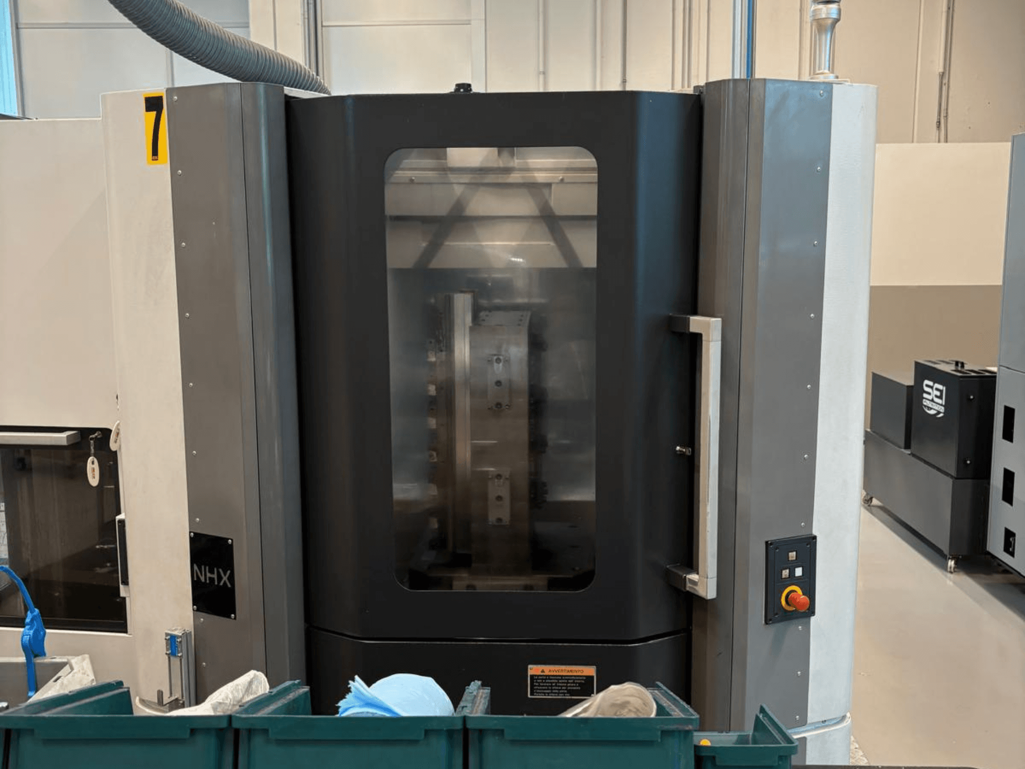 NHX CNC-kone edestä katsottuna, jossa näkyy ohjauspaneeli ja selkeä näyttö; sijaitsee työpajassa, jossa on vihreitä säiliöitä lähellä.