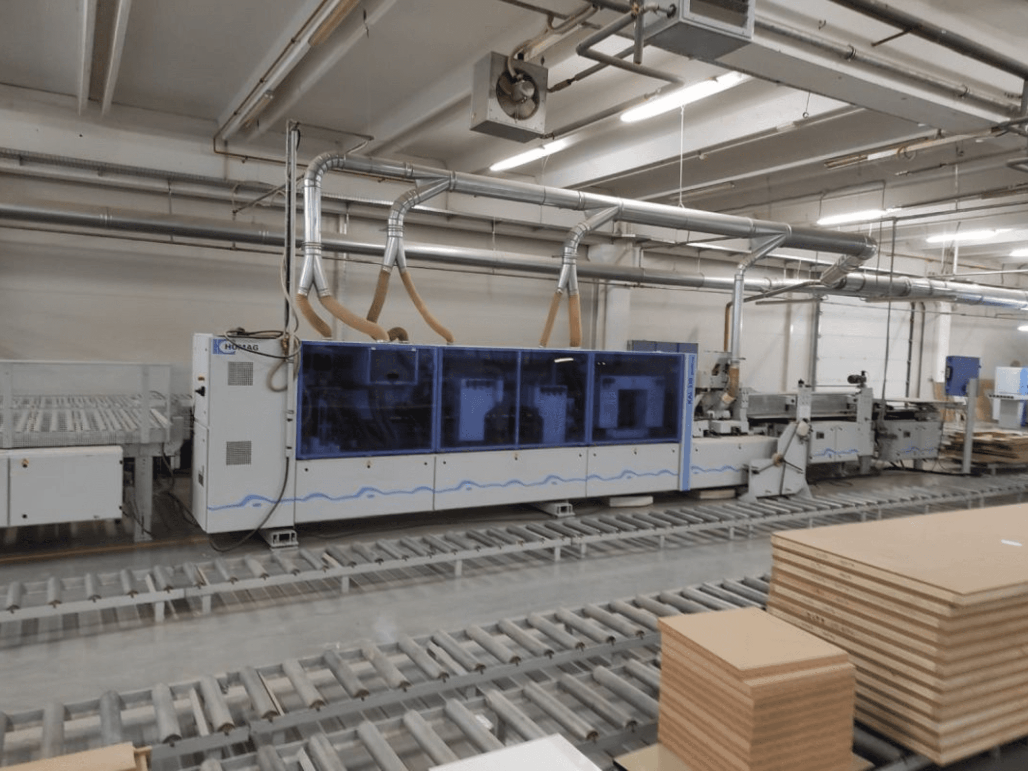 HOMAG-tuotantokone tehtaalla, etukuva, jossa sininen kotelo ja kuljetinjärjestelmä, jossa on pinottuja vanerilevyjä.