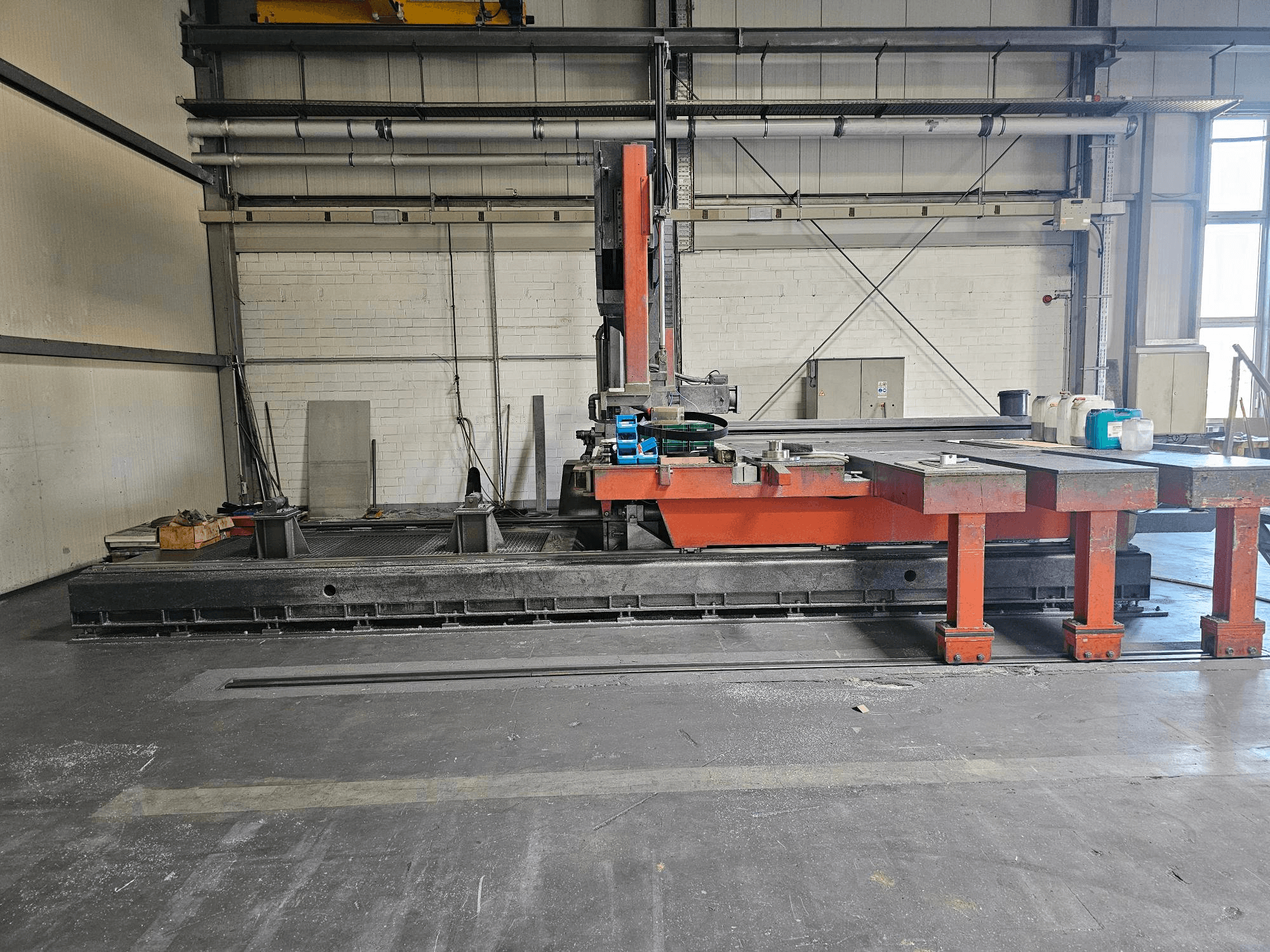 Teollisuusleikkauskone avoimessa työpajassa, oranssi ja musta, jossa on suuri pöytä; sivukuva, jossa näkyy lisävarusteet ja työtila.