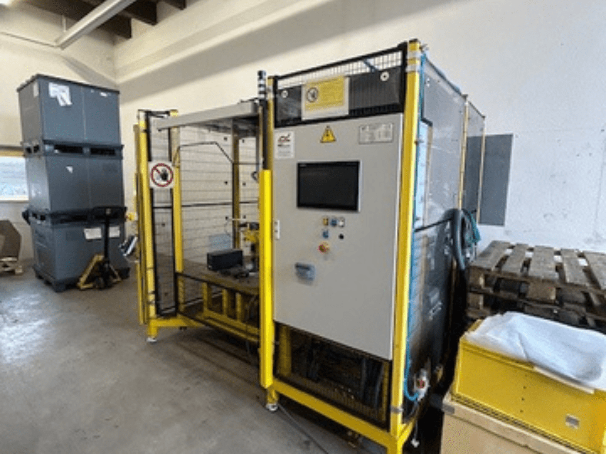 Teollisuuskone, jossa on ohjauspaneeli ja turvalaitteita, etukuva; taustalla keltainen kehys, harmaat säiliöt ja kuormalavat.