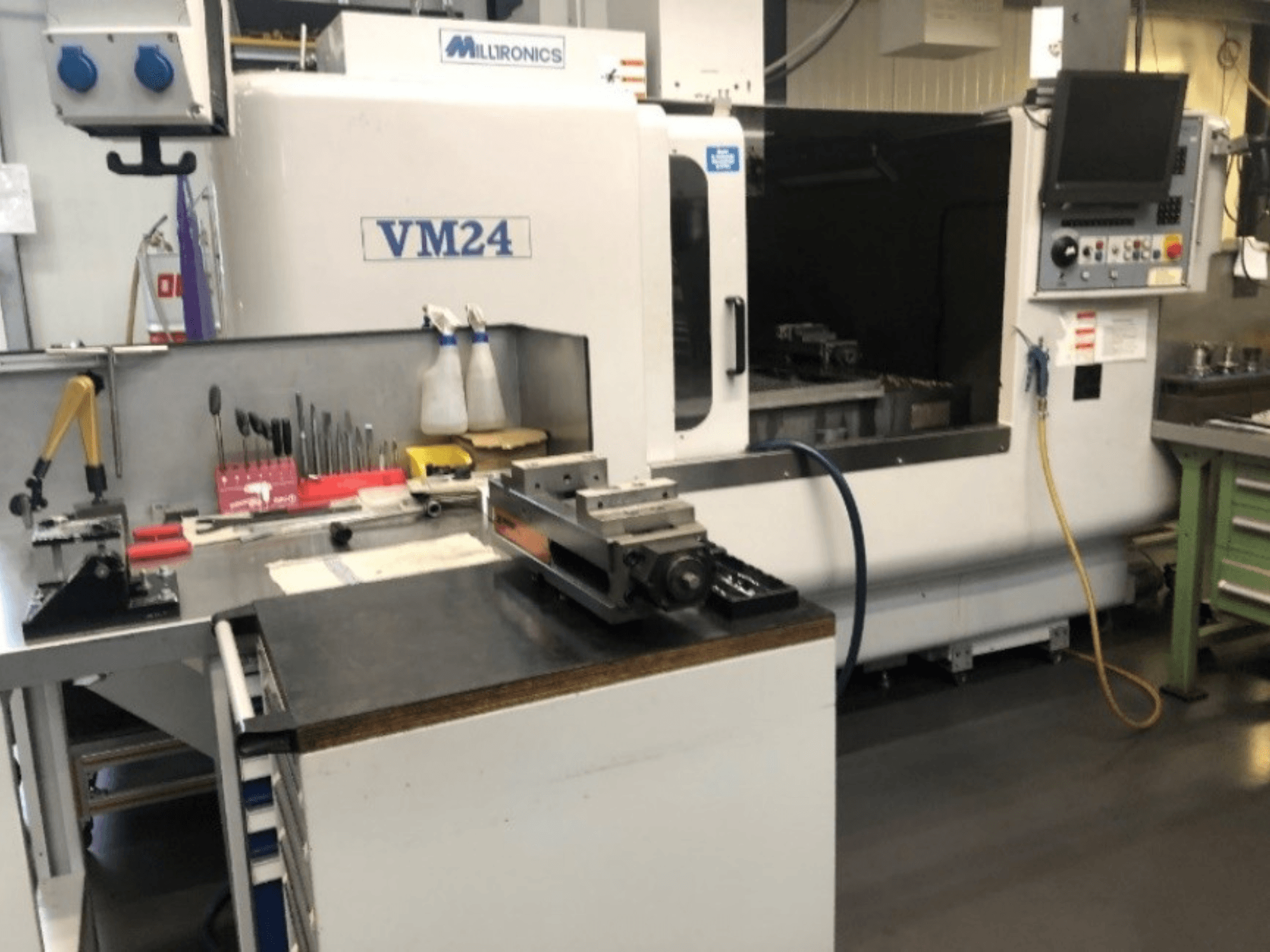Koneiden työtila, jossa on Milltronics VM24 CNC -kone. Etunäkymä, jossa työkaluja ja laitteita työpöydällä.
