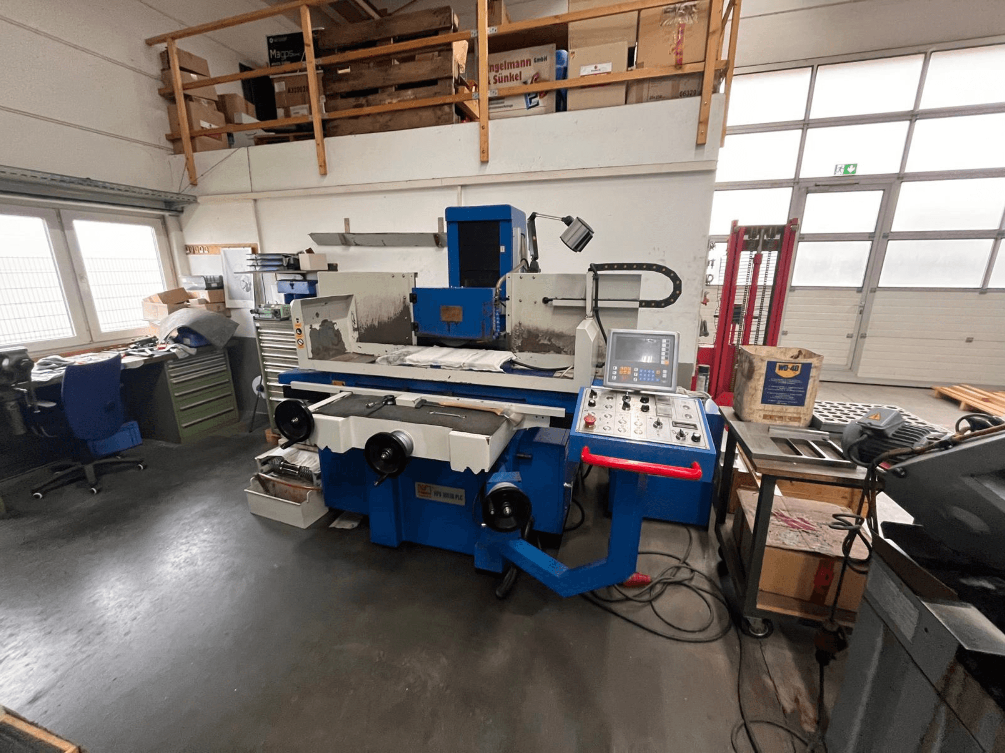 Sininen pintahiomakone, jossa on digitaalinen ohjauspaneeli, etukuva, jossa näkyy työtilan epäjärjestys ja vihreä työkalukaappi.