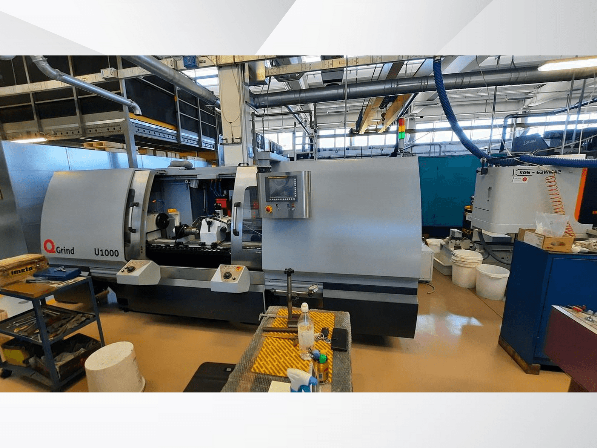 Q-Grind U1000 -kone työpajassa, etunäkymä, jossa on ohjauspaneeli ja avoin leikkausalue, jota ympäröivät laitteet.