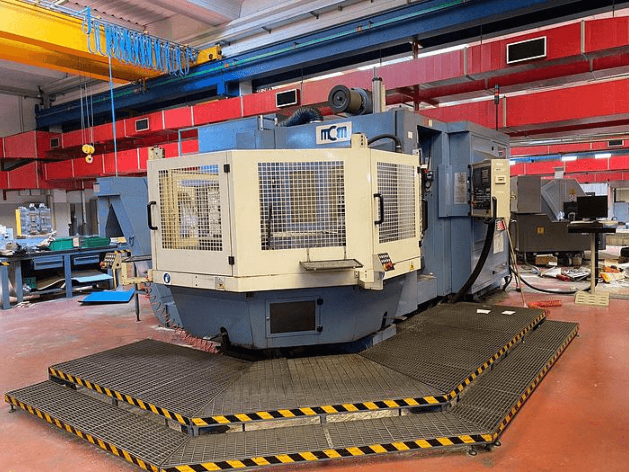 MCM CNC-kone tehtaalla, etukuva suojahäkillä, mustat ja keltaiset turvareunat, teollisuuslaitteiden ympäröimänä.