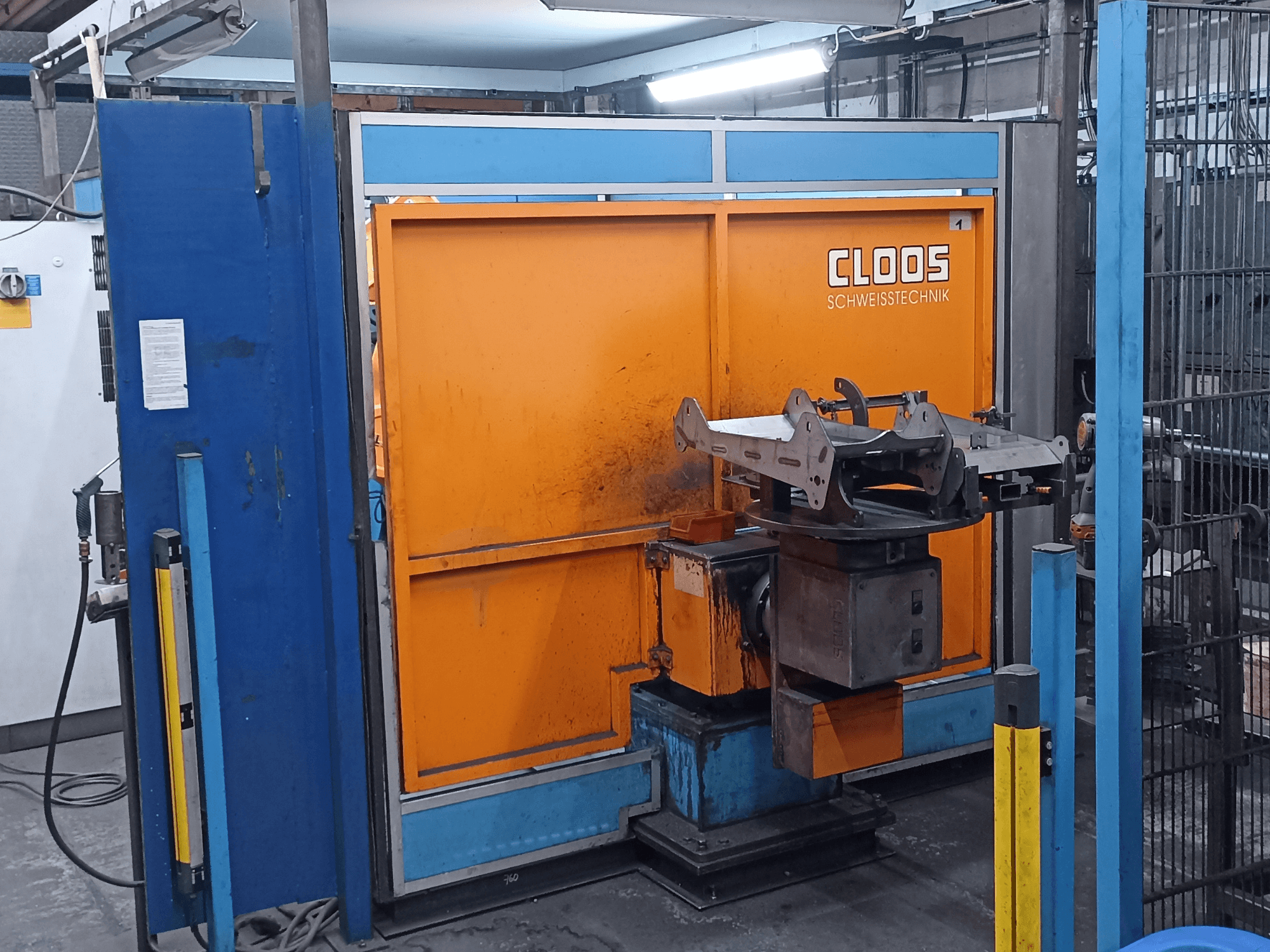 CLOOS Romat 320 -hitsausrobotti työpajassa, edestä katsottuna, oranssilla ohjauspaneelilla ja metallisella työkappaleella pyörivällä alustalla.