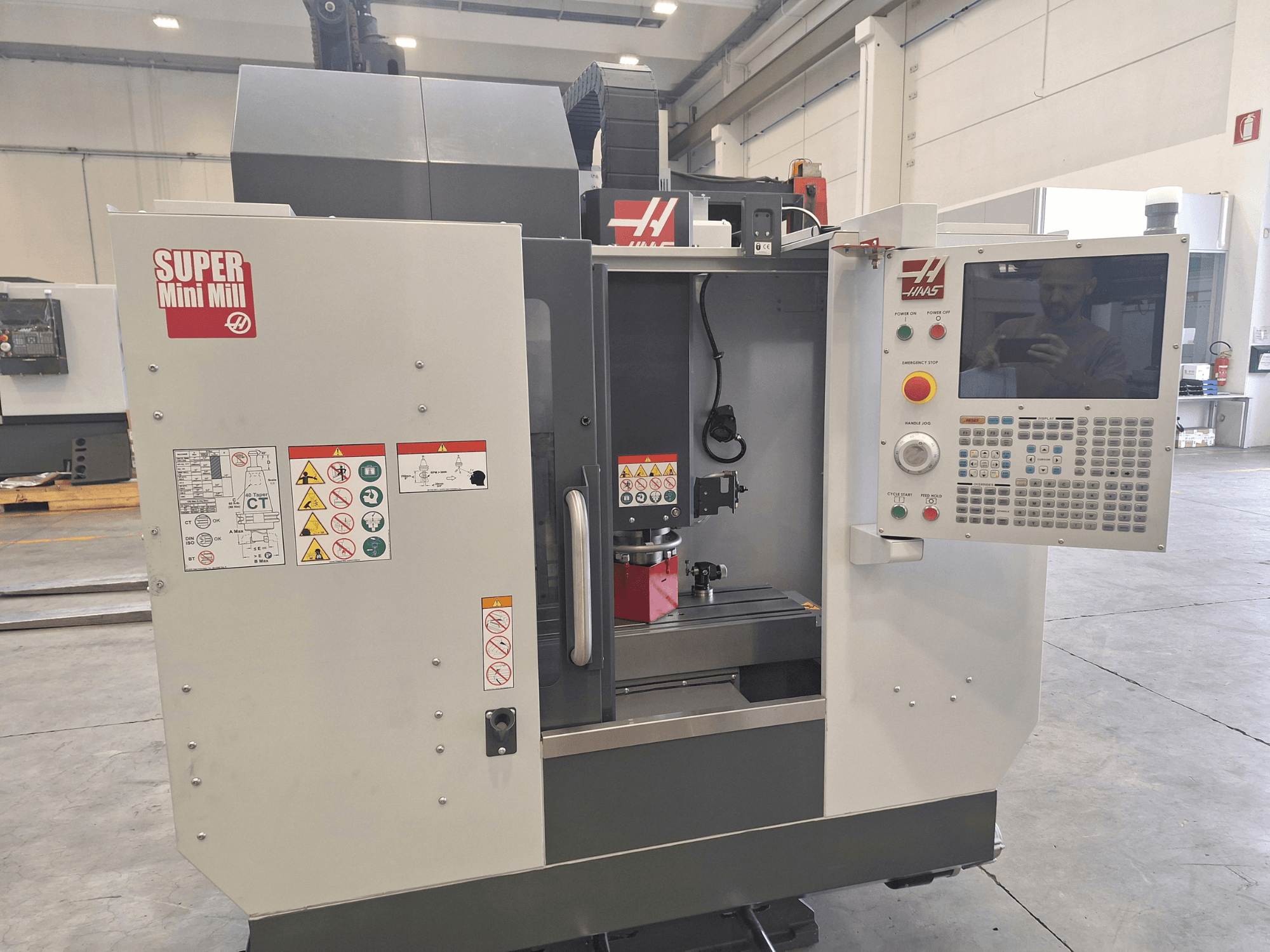 HAAS Super Mini Mill -koneen etunäkymä, jossa näkyvät ohjauspaneeli, turvallisuusmerkinnät ja työstöalue, jolla on työkappale asennettuna.