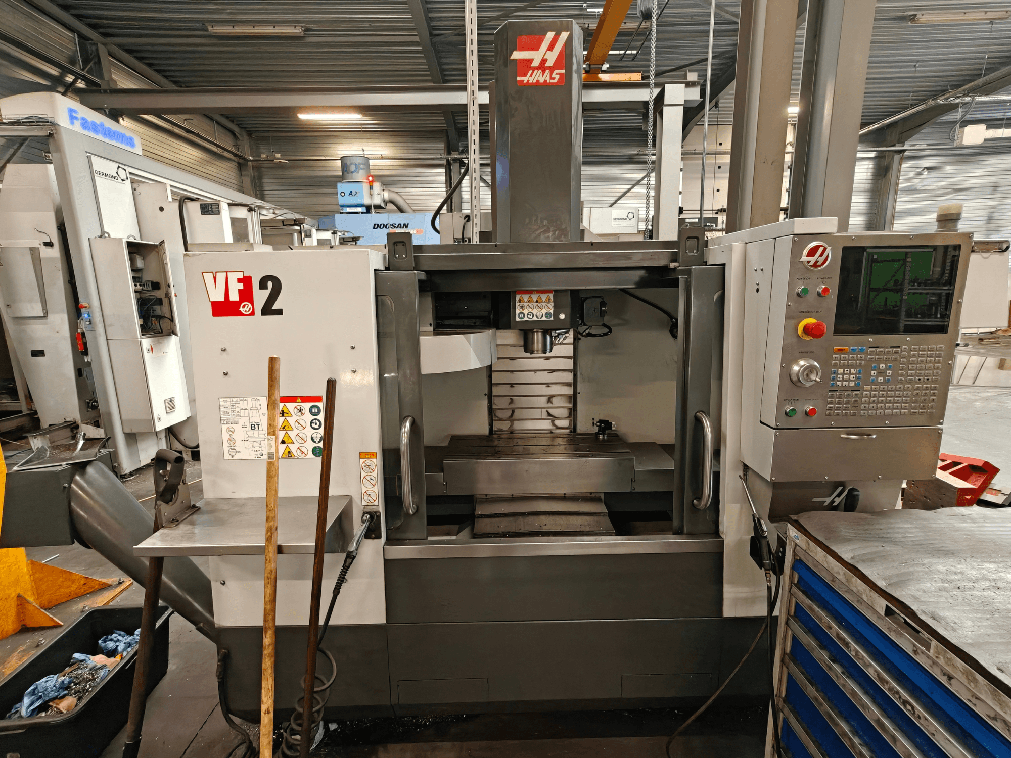 HAAS VF-2 -pystykoneistuskeskus, etunäkymä, jossa näkyvät ohjauslaitteet, kara ja työpöytä työpajaympäristössä.