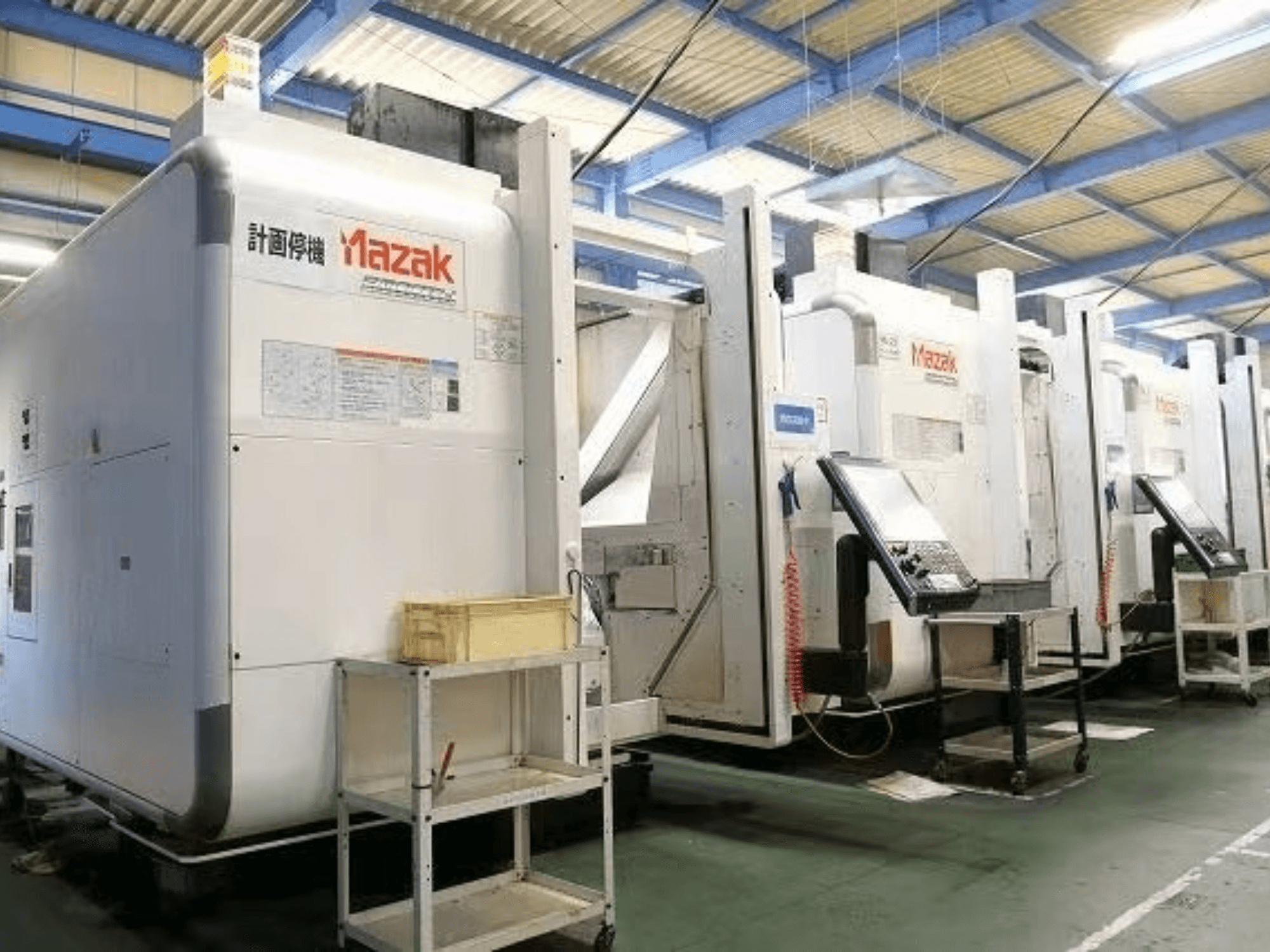 Mazak CNC-koneet tehdasympäristössä, sivukuva, jossa näkyy ohjauspaneelit ja tarrat valkoisilla rungoilla.