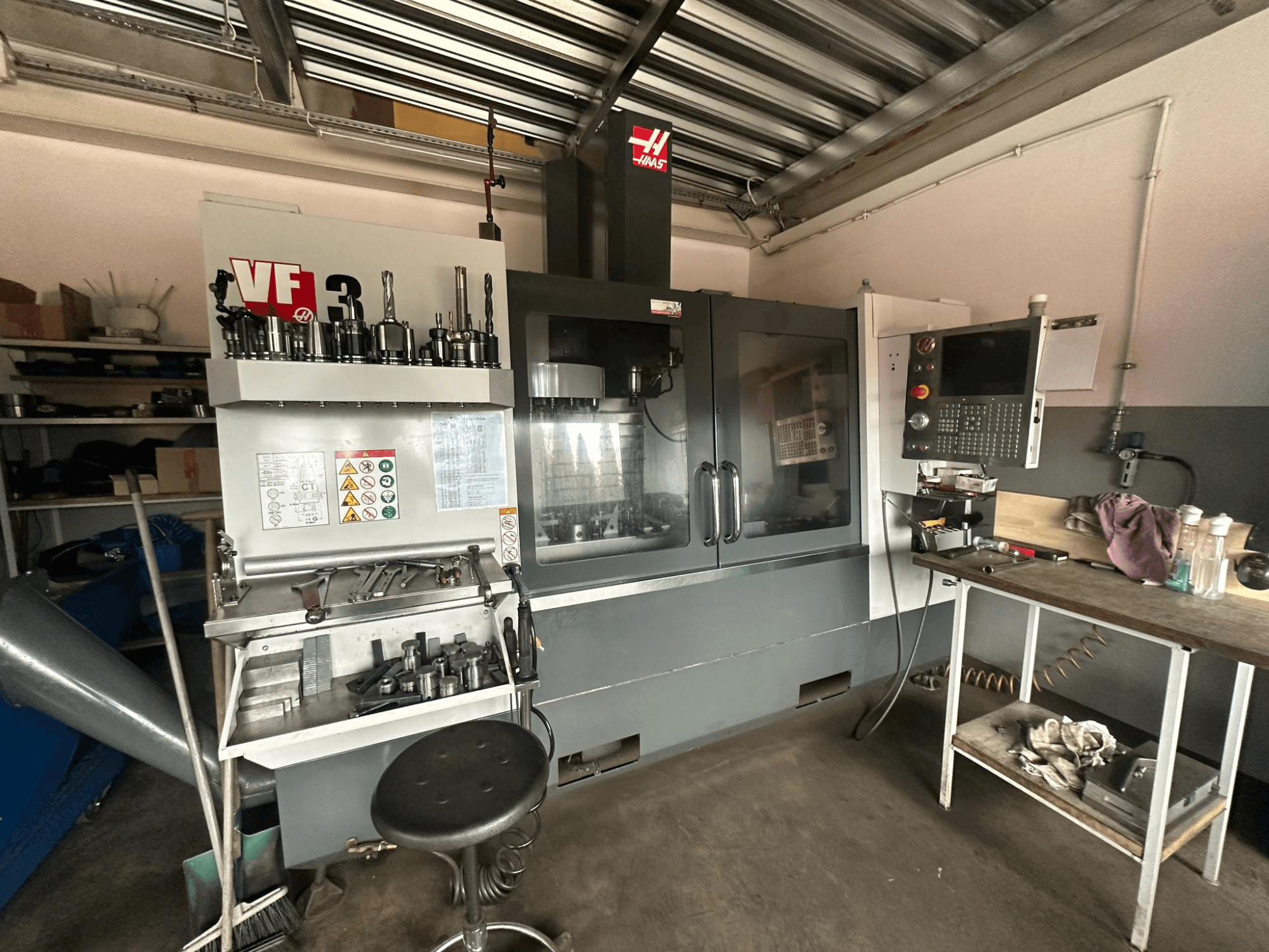 Haas VF-3 CNC-kone työpajassa, etukuva, työkalut ja hallintalaitteet viereisellä pöydällä, tekninen käyttöliittymä ja turvallisuustiedot.
