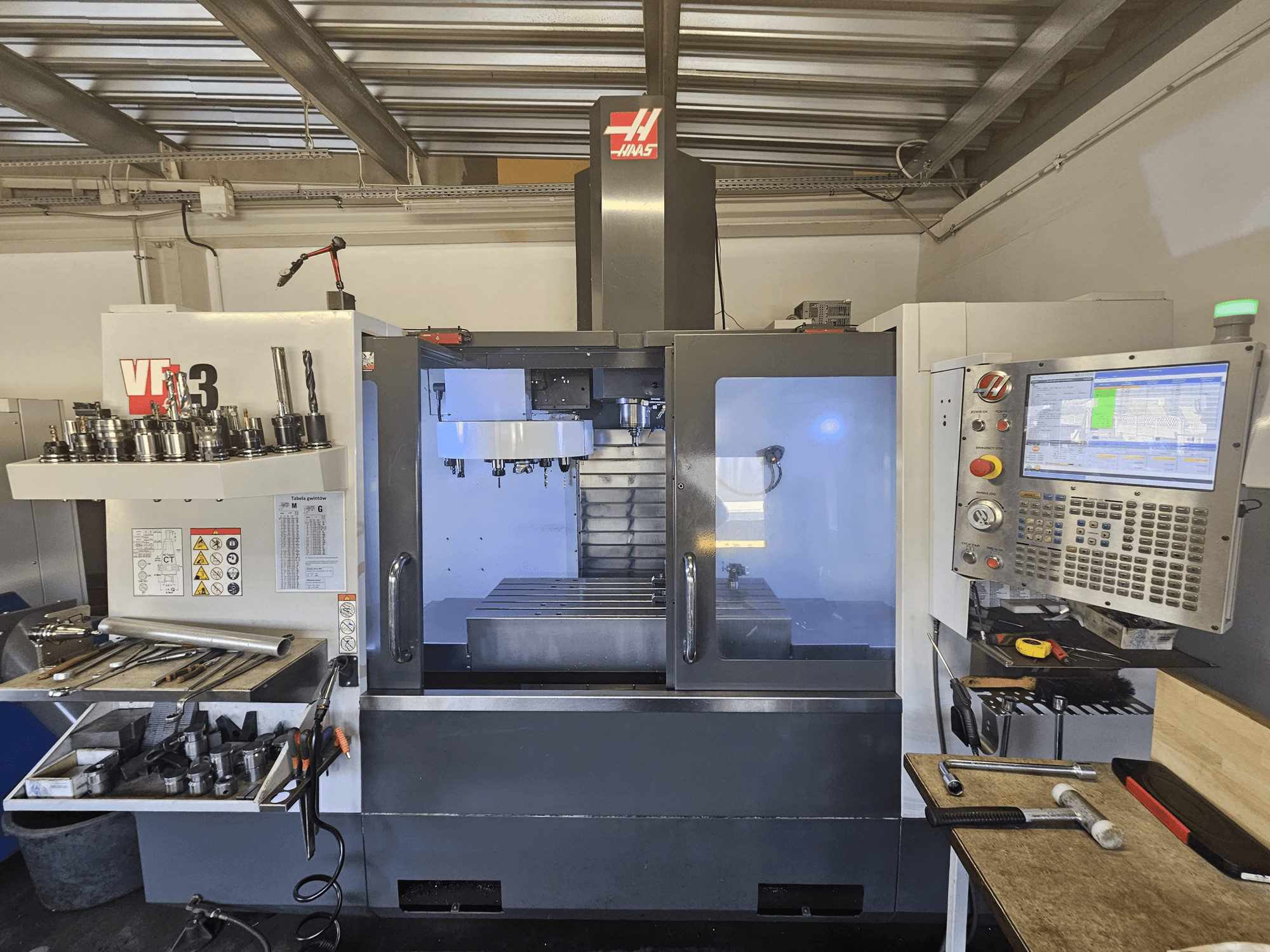 Haas VF-3 CNC-kone työpajassa, etunäkymä, jossa näkyvät työkalut, hallintalaitteet ja jyrsintäalue valaistulla sisätilalla.