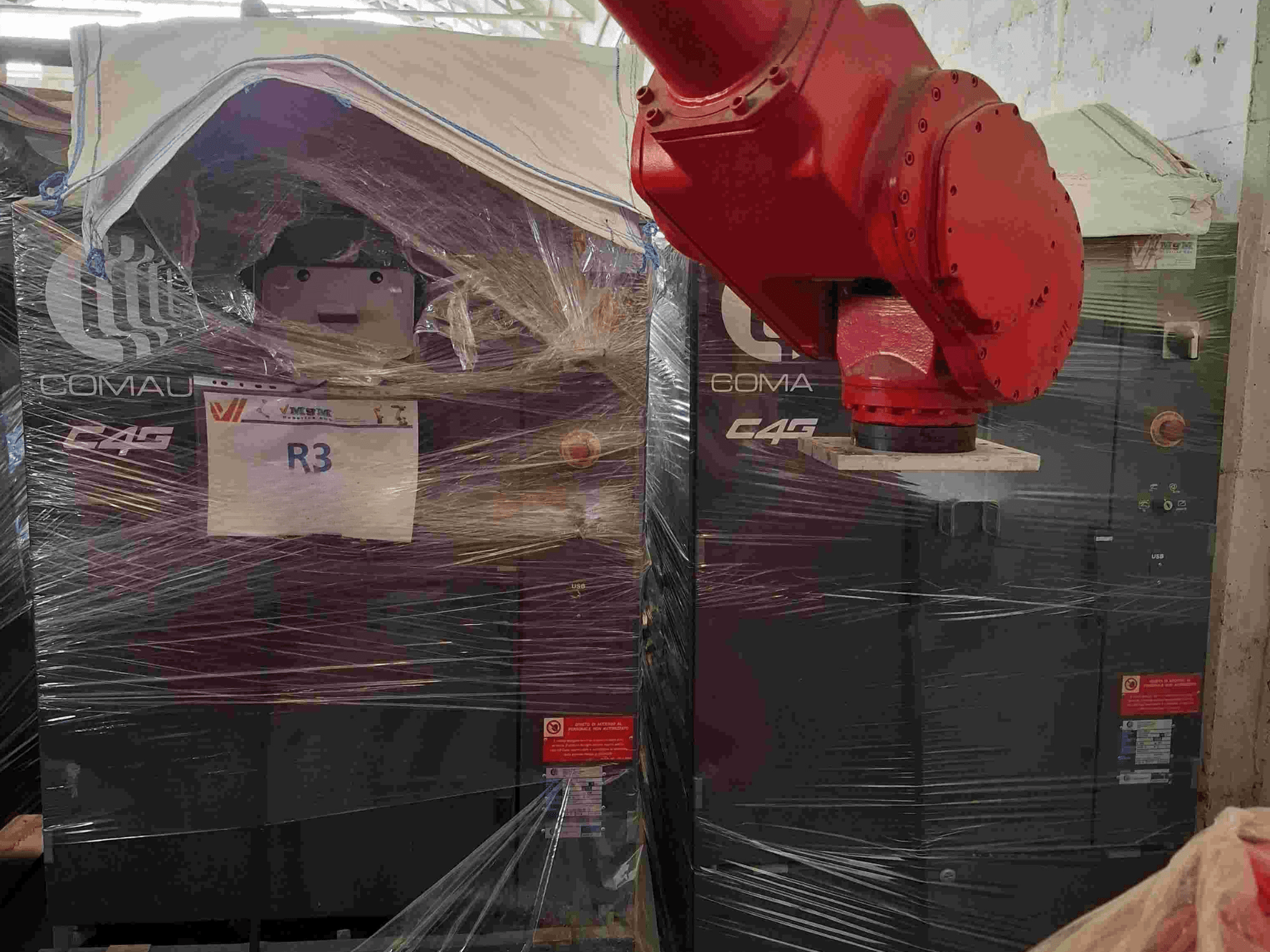 Comau C4G -robottikäsivarsi edestä katsottuna, muoviin käärittynä, merkinnällä "R3" ja koneessa näkyvillä varoitustarroilla.