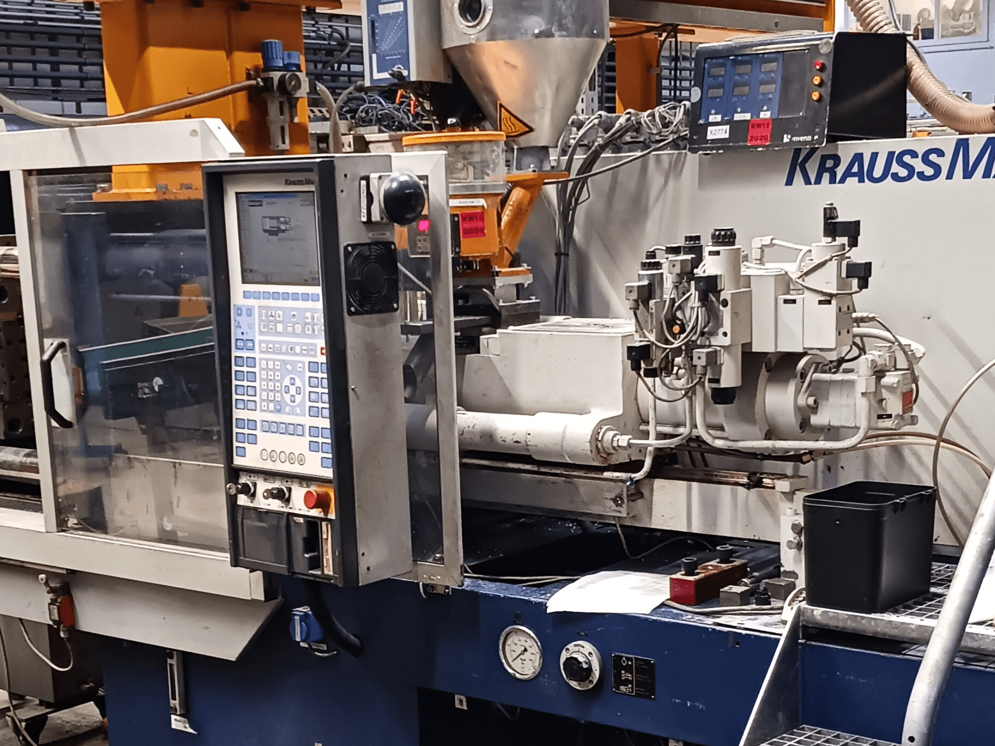 KraussMaffein ruiskuvalukone, jossa on ohjauspaneeli, hydrauliikkayksiköt ja työkalut etualalla, etukuva.