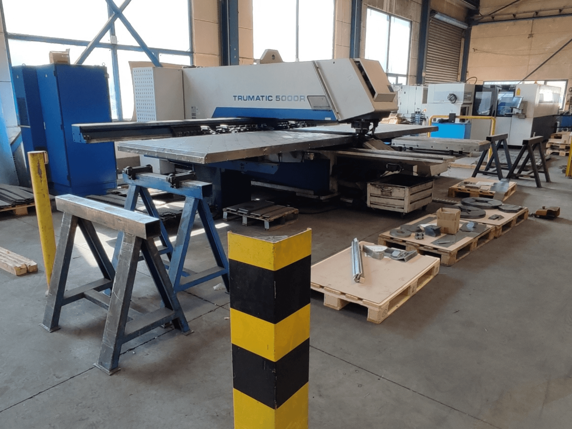 TRUMPF Trumatic 5000 R -CNC-lävistyskone työpajassa, sivukuva, jonka vieressä on työkaluja ja materiaaleja kuormalavoilla.