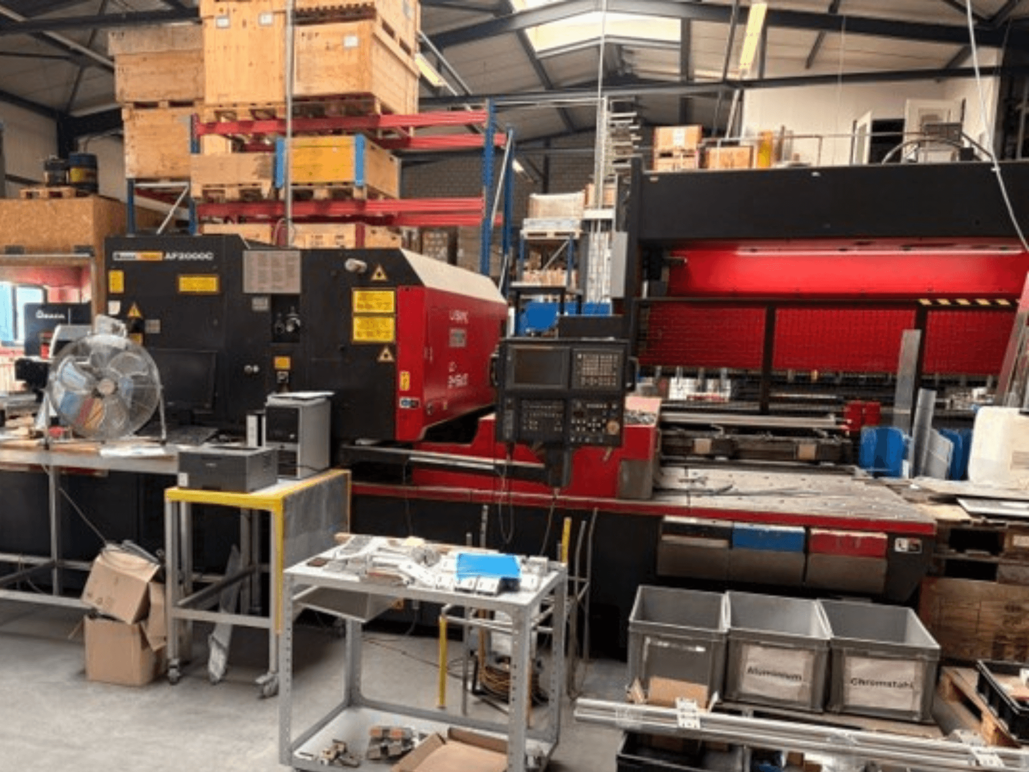 AMADA LC-2415αIII CO2-laserleikkauskone työpajaympäristössä; kuvassa näkyy etunäkymä, ohjauspaneeli ja työskentelytila.