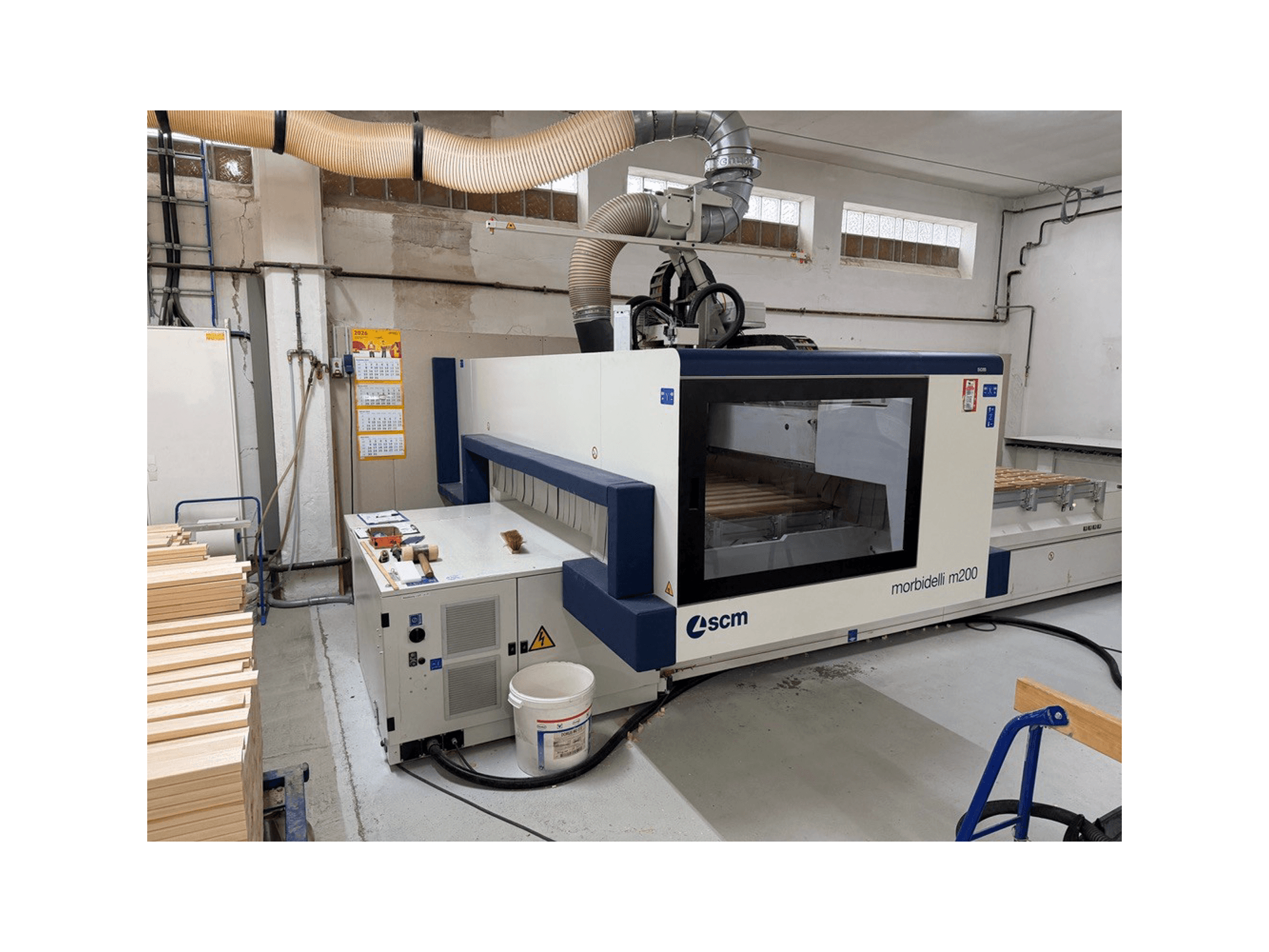 SCM Morbidelli M200F CNC-työstökeskus työpajassa, sivukuva, jossa pöydällä on puukappaleita ja yläpuolella ilmanvaihtojärjestelmä.