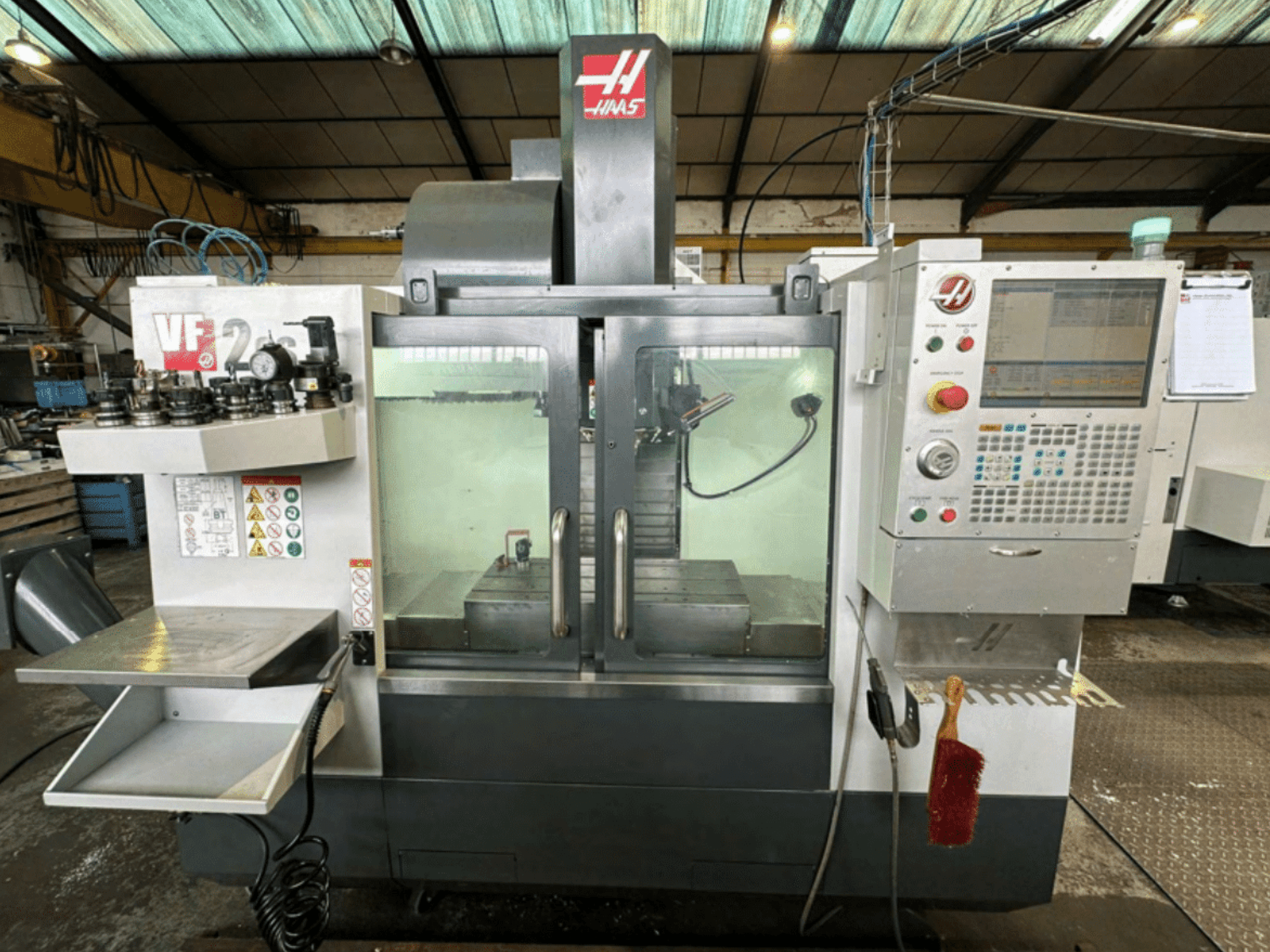 Haas VF2 CNC-koneen etukuva, jossa näkyy ohjauspaneeli, työkalunpitimet ja työtila, jossa on turvatarroja ja harja.