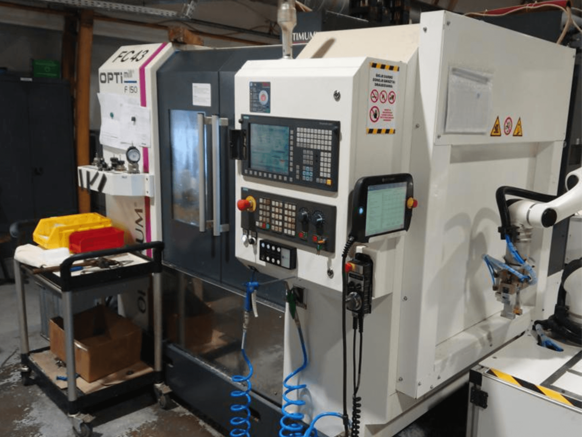 OPTIMUM F50 -jyrsinkoneen ohjauspaneeli, jossa on letkuja, työkaluja ja robottikäsivarsi näkyvissä työpajassa.