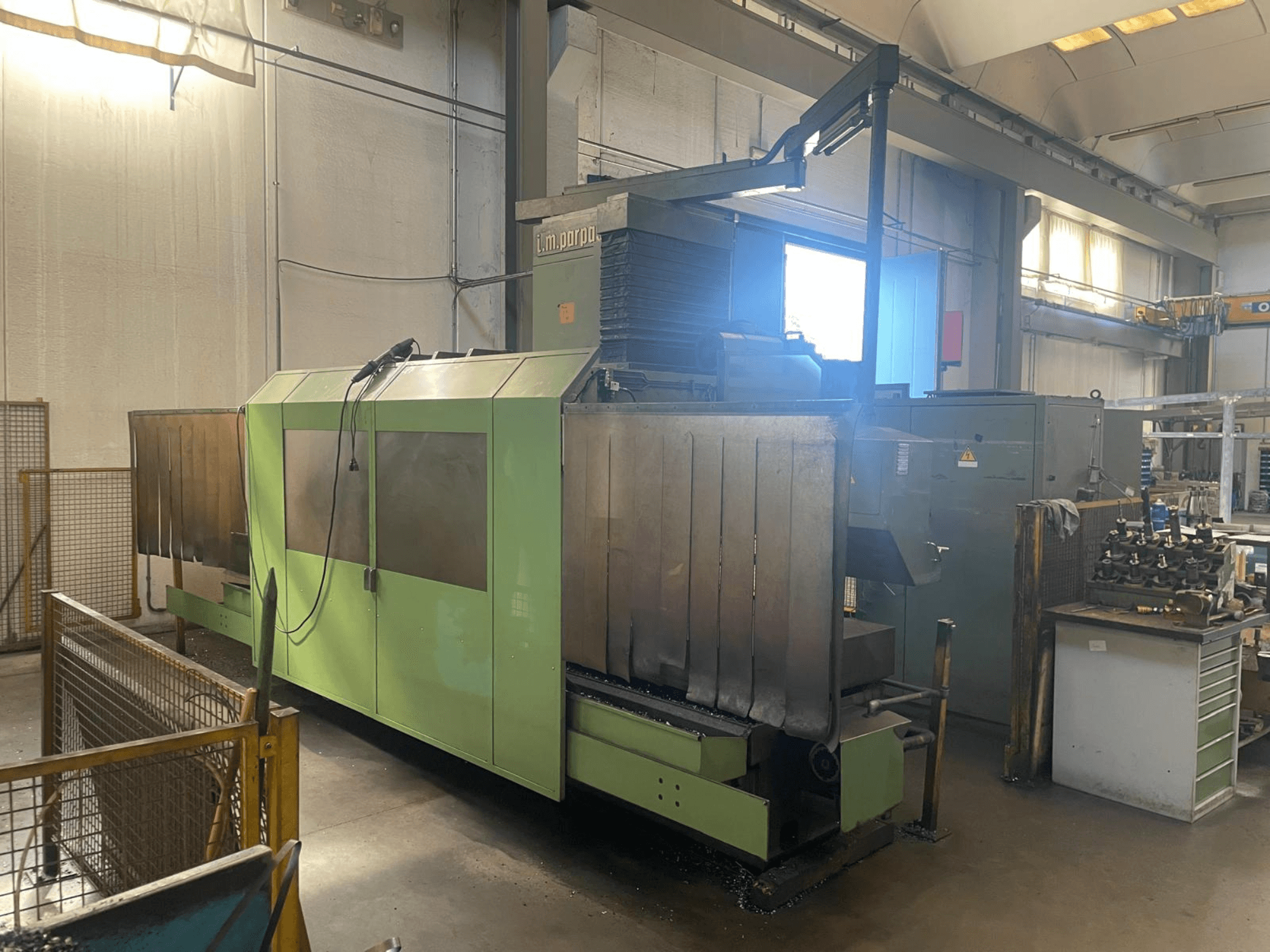 Vihreä teollisuuskone, jossa on näkyviä metallikomponentteja, etukuva; siinä on ohjauspaneeli, turvavaroituksia ja työpiste lähellä.