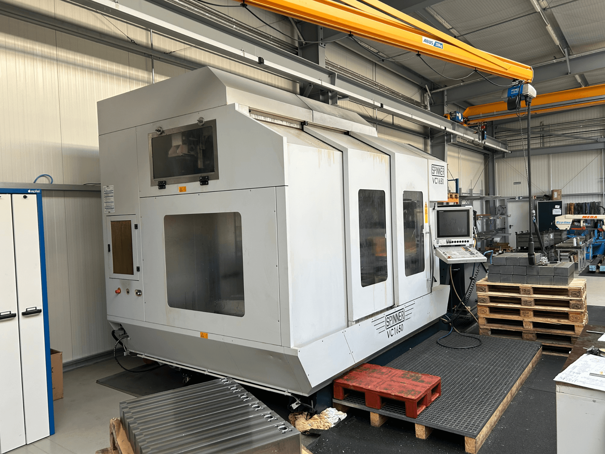 Spinner VC 1650 CNC-kone, etukuva, jossa on ohjauspaneeli, suuret ikkunat ja lähistöllä pinottuja kuormalavoja työpajassa.