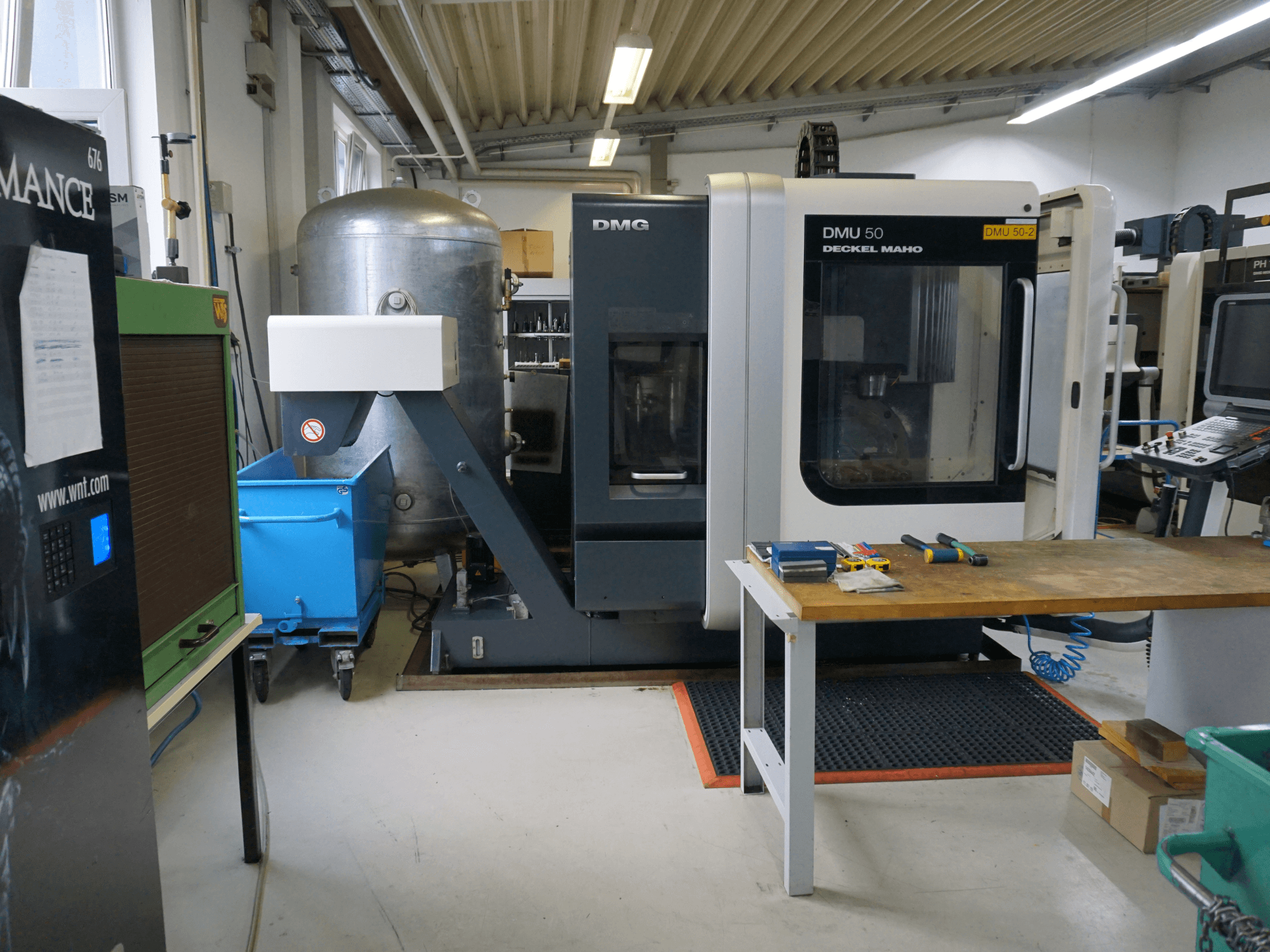 DMG DMU 50 -jyrsinkone työpajassa, etukuva, työkalut puupöydällä ja sininen pyörällinen roskakori vieressä.