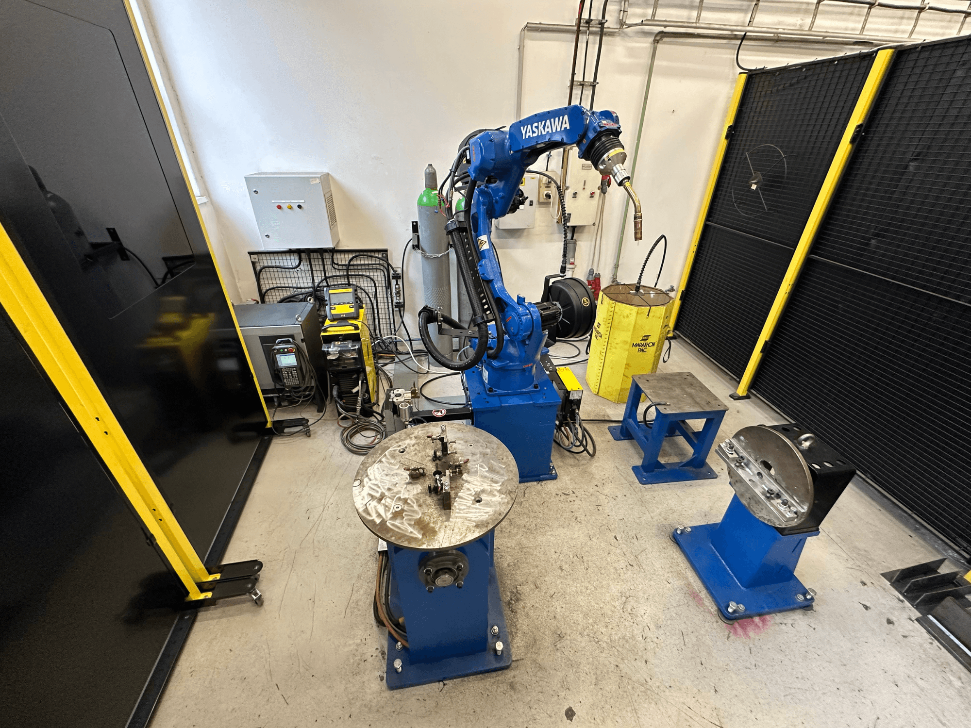 Sininen Yaskawa-robottikäsivarsi, kaksi työpöytää, hitsauslaitteet ja turvakaiteet; teollinen työtila edestäpäin.