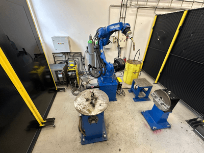 Sininen Yaskawa-robottikäsivarsi, kaksi työpöytää, hitsauslaitteet ja turvakaiteet; teollinen työtila edestäpäin.
