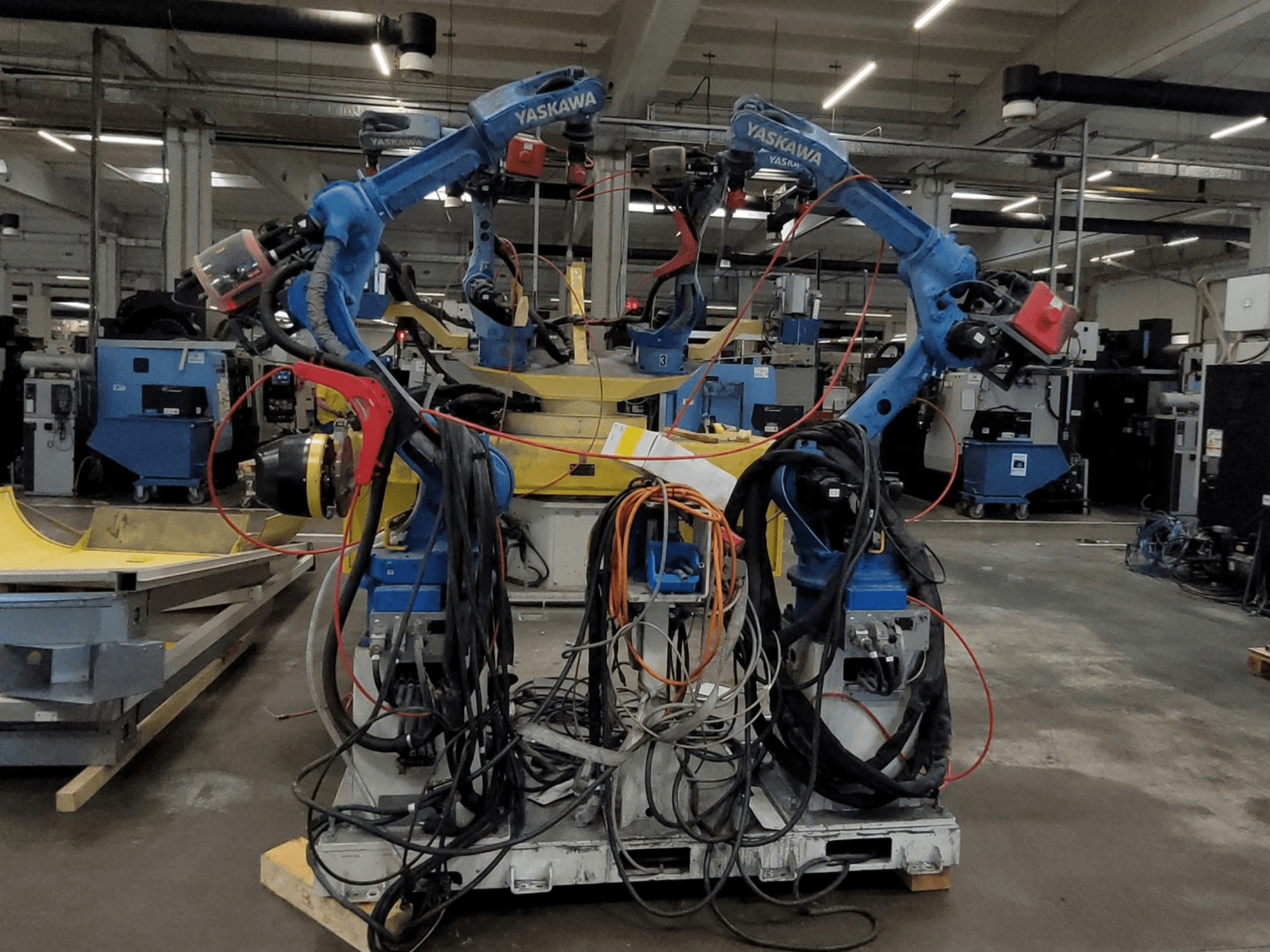 Yaskawan robottihitsausyksikkö RWC L1_OS 15330, etunäkymä, jossa on kaksi sinistä robottia sekä runsaasti kaapeleita ja hitsaustyökaluja.