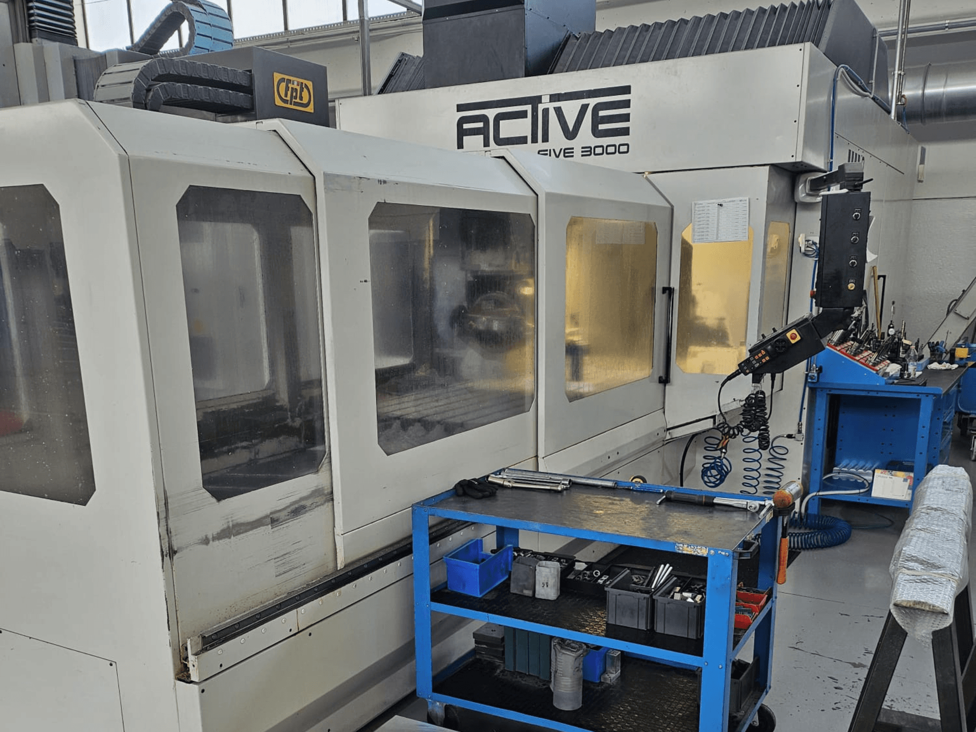 Aktiivinen 3000 CNC-kone työpajassa, etunäkymä, työkalut sinisellä kärryllä, näkyvissä olevat hallintalaitteet ja työalue sisällä.