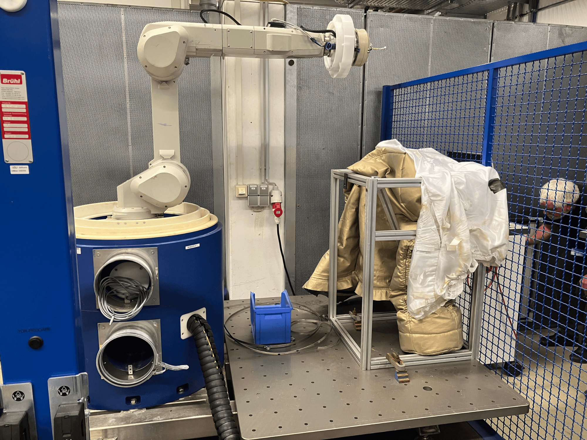 Robottikäsi lähellä sinistä kammiota, jossa on valkoinen ja kultainen puku metallisella kehyksellä, sivukuva turvaverkkotaustalla.