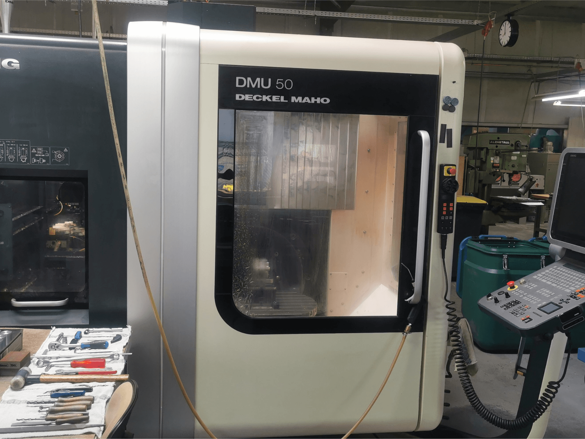 DMU 50 deckel maho CNC-kone edestä katsottuna, jossa näkyy ohjauspaneeli ja läpinäkyvä ovi. Työkalut ovat lähellä.
