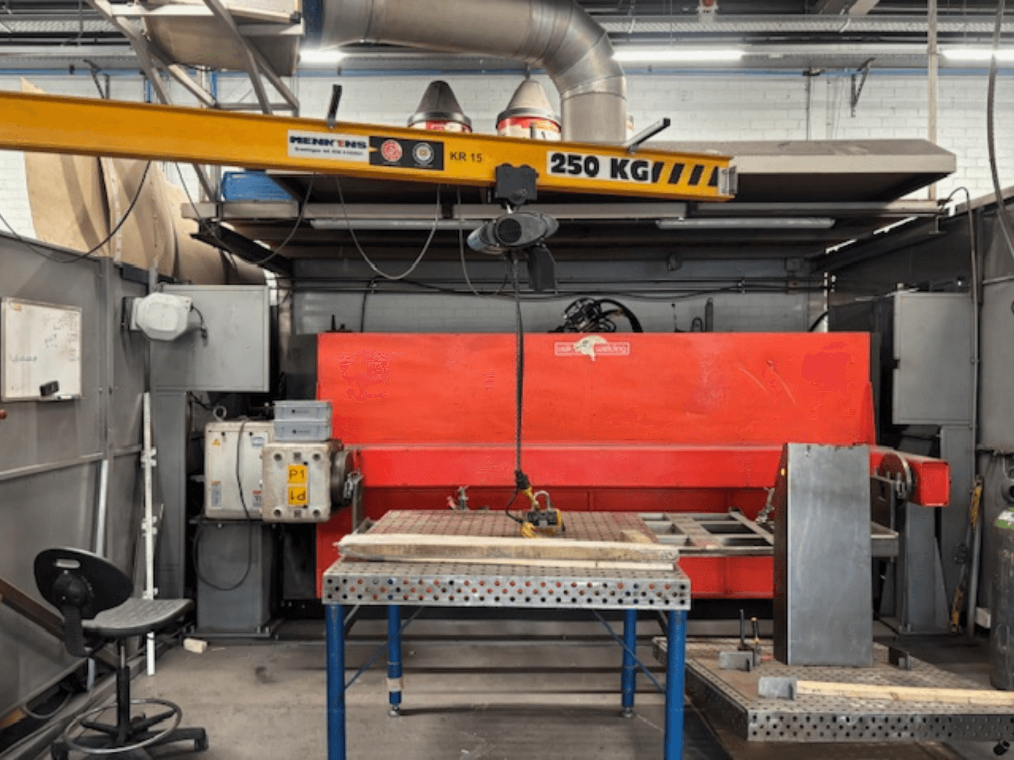 Teollisuustyöpaja, jossa on punainen hydraulinen puristin, nosturi, jossa on merkintä "KR 15", ja metallinen työpöytä, sivuperspektiivi.