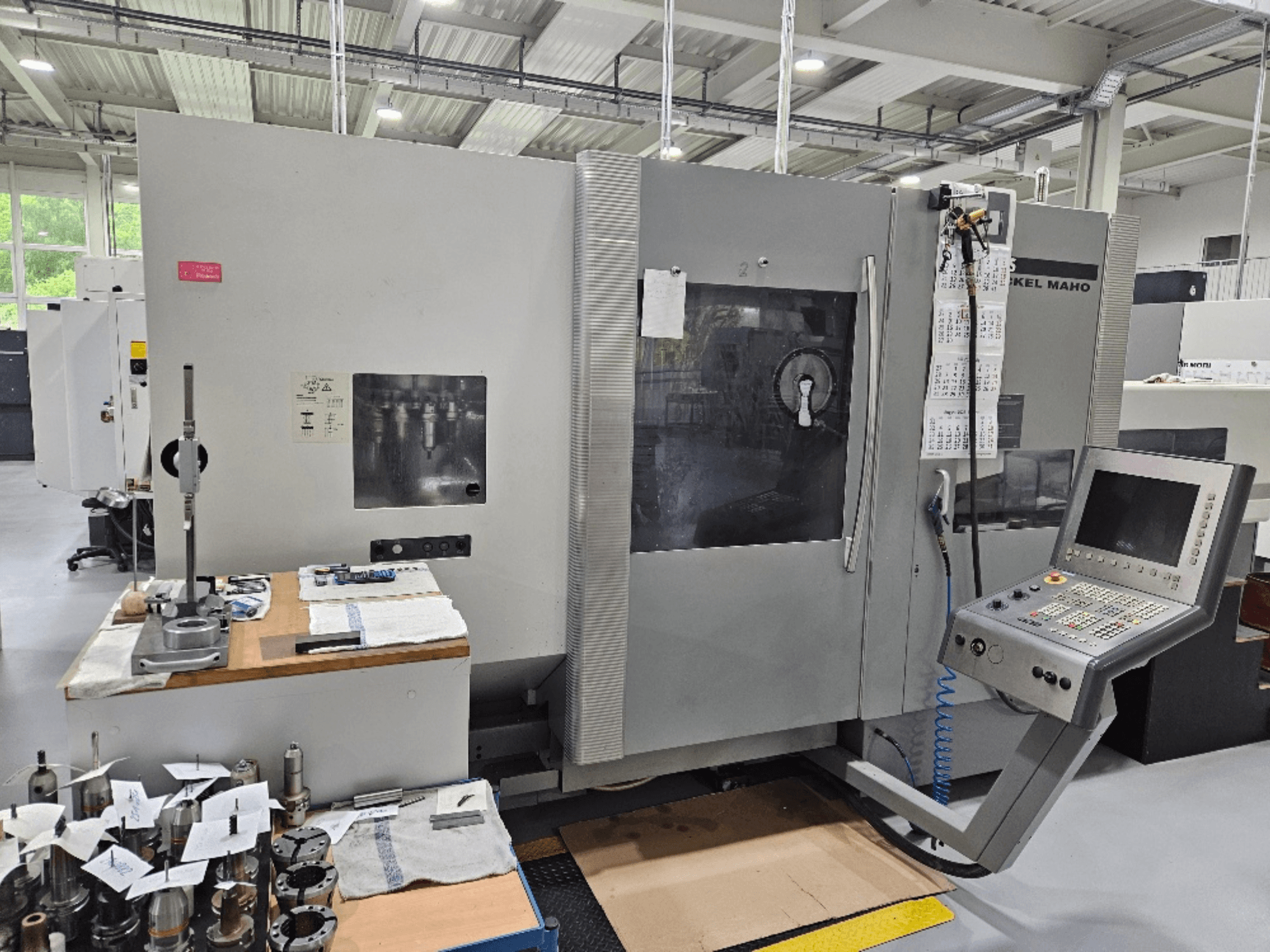 Etukuva DMG Mori DMU 70 -koneesta, jossa on ohjauspaneeli, työkaluja puupöydällä ja erilaisia työkappaleita lähellä.