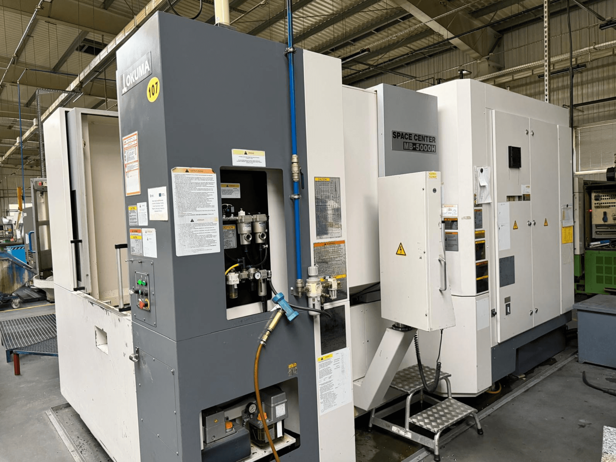 Okuma MB 5000H -vaakakoneistuskeskus, sivukuva, jossa näkyy ohjauspaneeli ja koneen osat tehdasympäristössä.