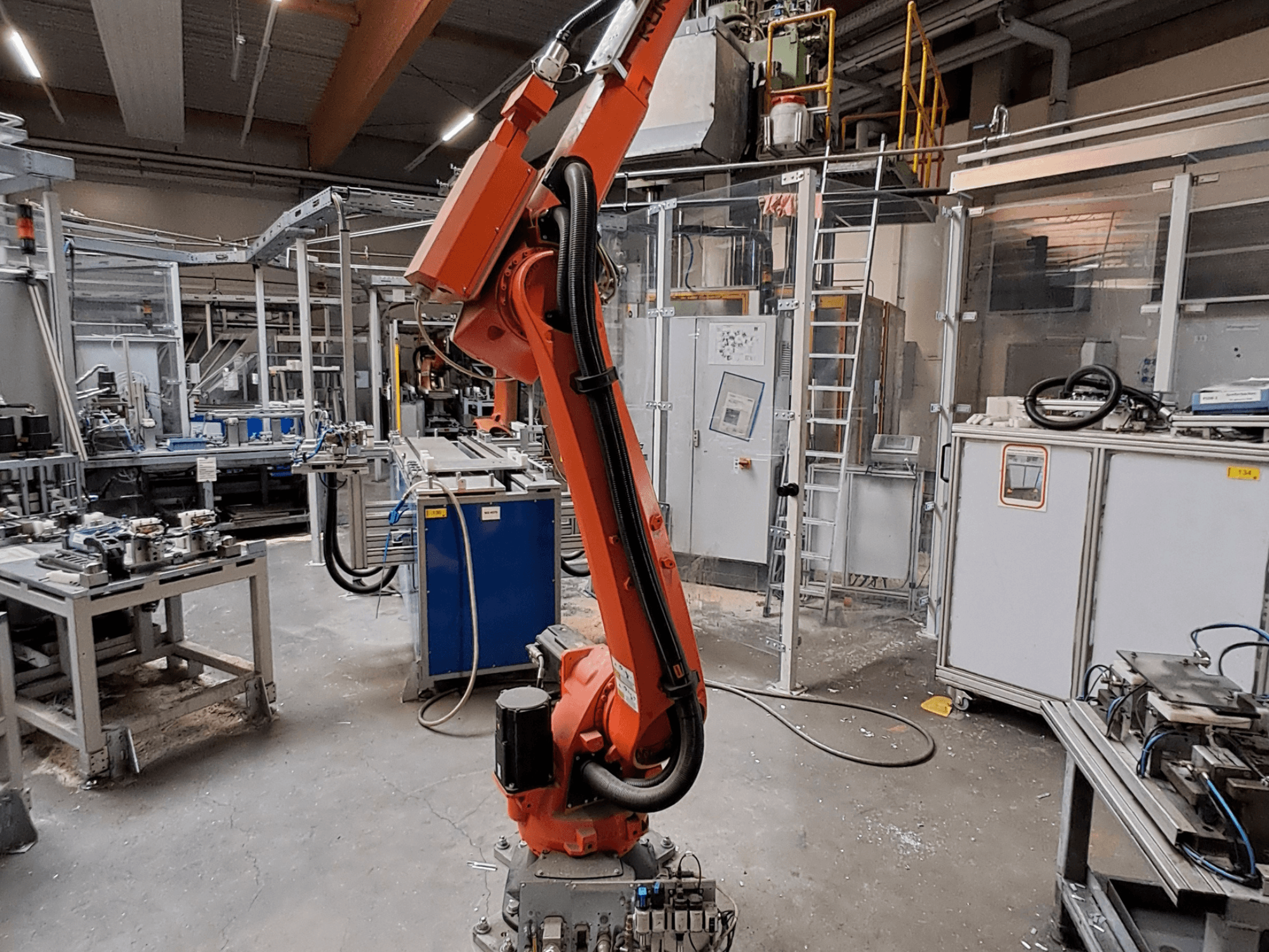 KUKA KR 26 R3100/SEL -robottivarsi teollisuusympäristössä, sivulta kuvattuna, taustalla toiminnassa olevia laitteita.