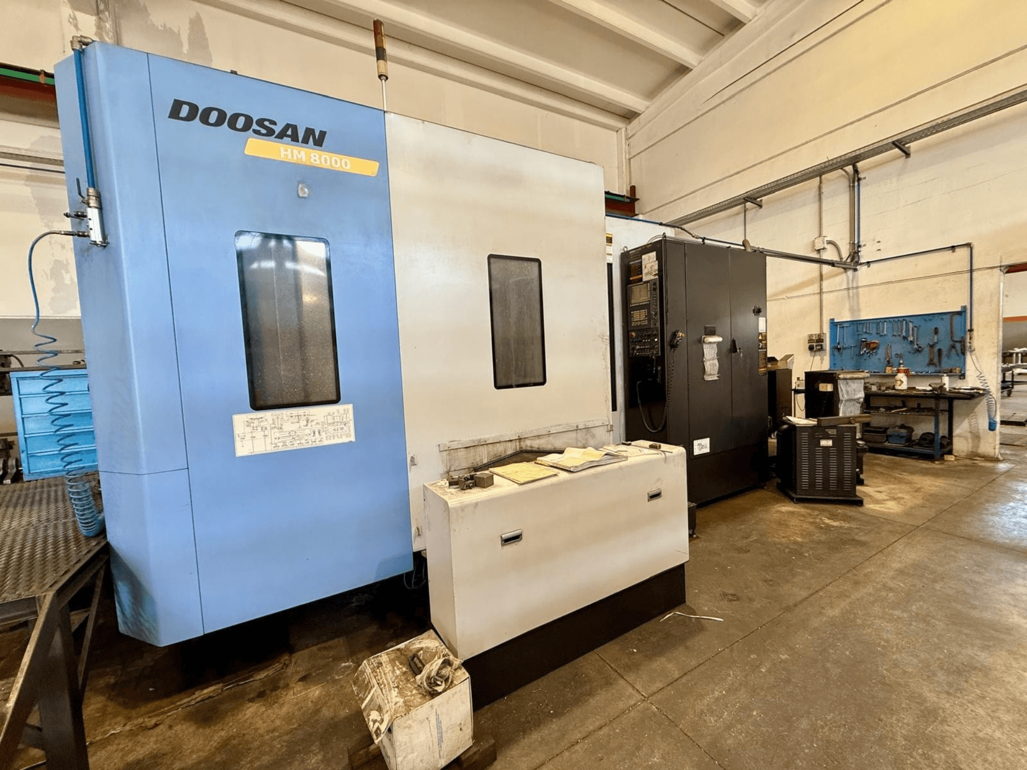 Sininen DOOSAN HM 8000 -vaakakoneistuskeskus, etunäkymä, jossa näkyvät ohjauspaneeli ja sen vieressä olevat työpajavälineet.