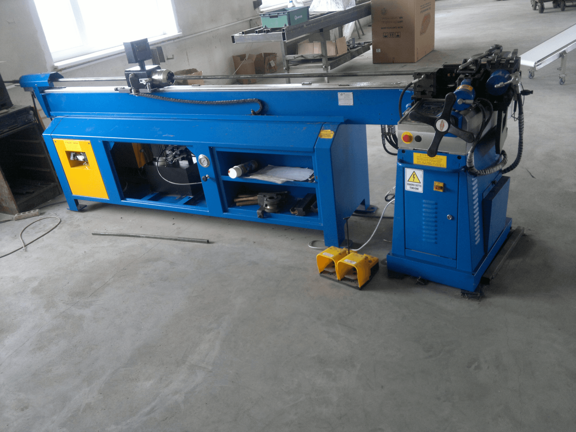 Sininen puuntyöstökone keltaisilla korostuksilla, sivukuva; siinä on ohjauspaneeli, jalkapolkimet ja pyörivä teräasennelma.