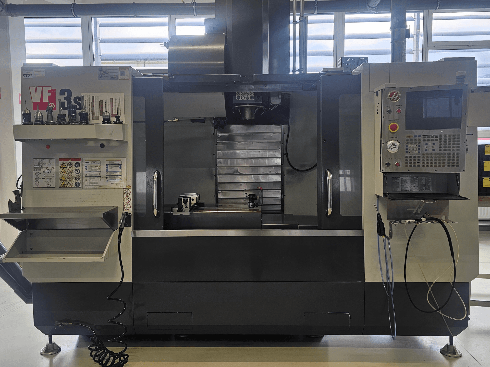 Haas VF3SS CNC-jyrsin, etukuva, jossa on työkalunpitimet, ohjauspaneeli ja tyhjä jäähdytysnestelokero, jossa on turvatarrat näkyvissä.