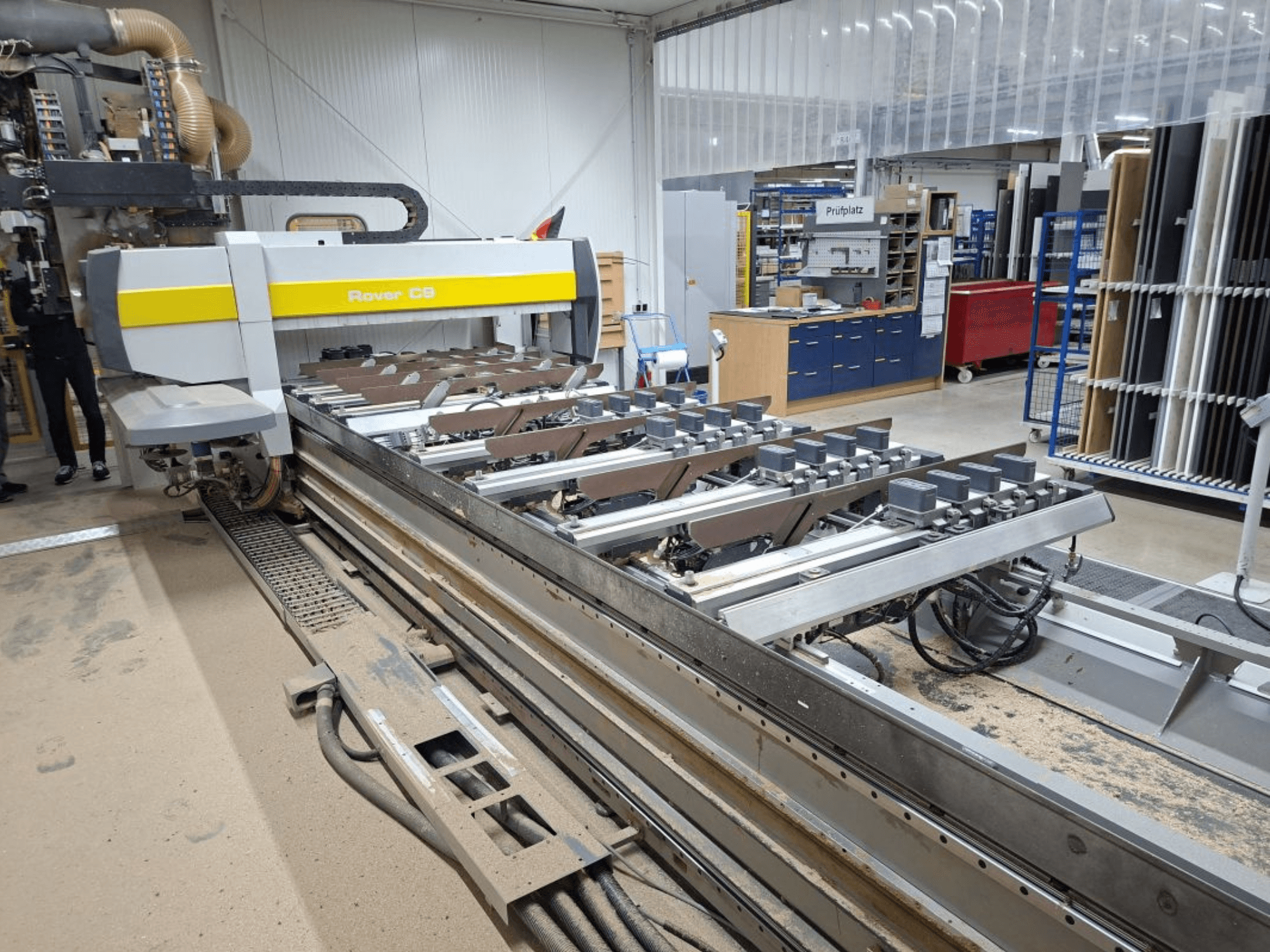 Rover C9 CNC-kone työpajassa; etukuvassa näkyvät leikkuutyökalut ja kuljetinjärjestelmä, ja lattialle on kertynyt pölyä.