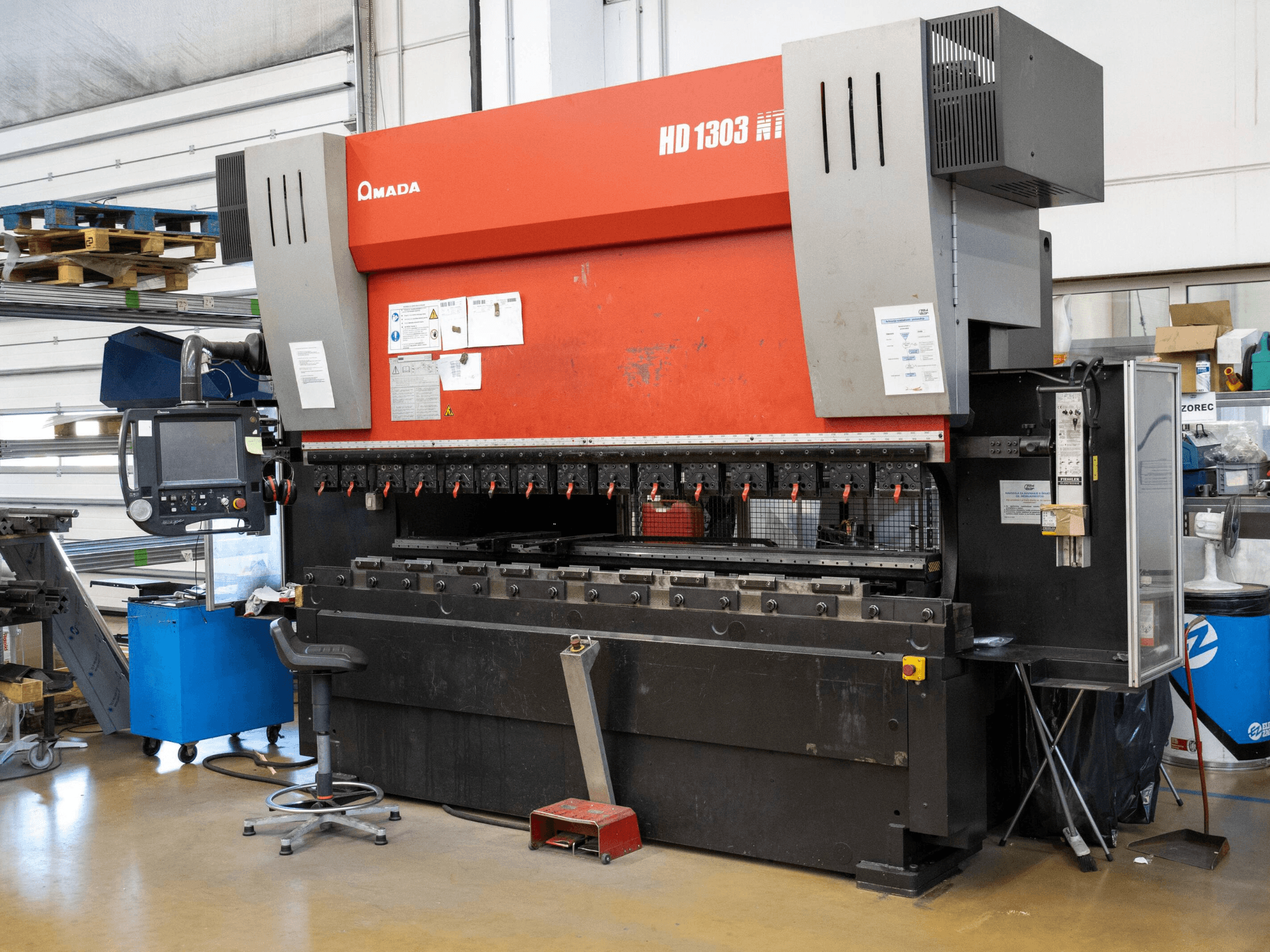 Amada HD 1303 hydraulinen puristuspuristin työpajassa, etukuva; siinä on useita puristimia, ohjauspaneeli ja säädettävä jakkara.
